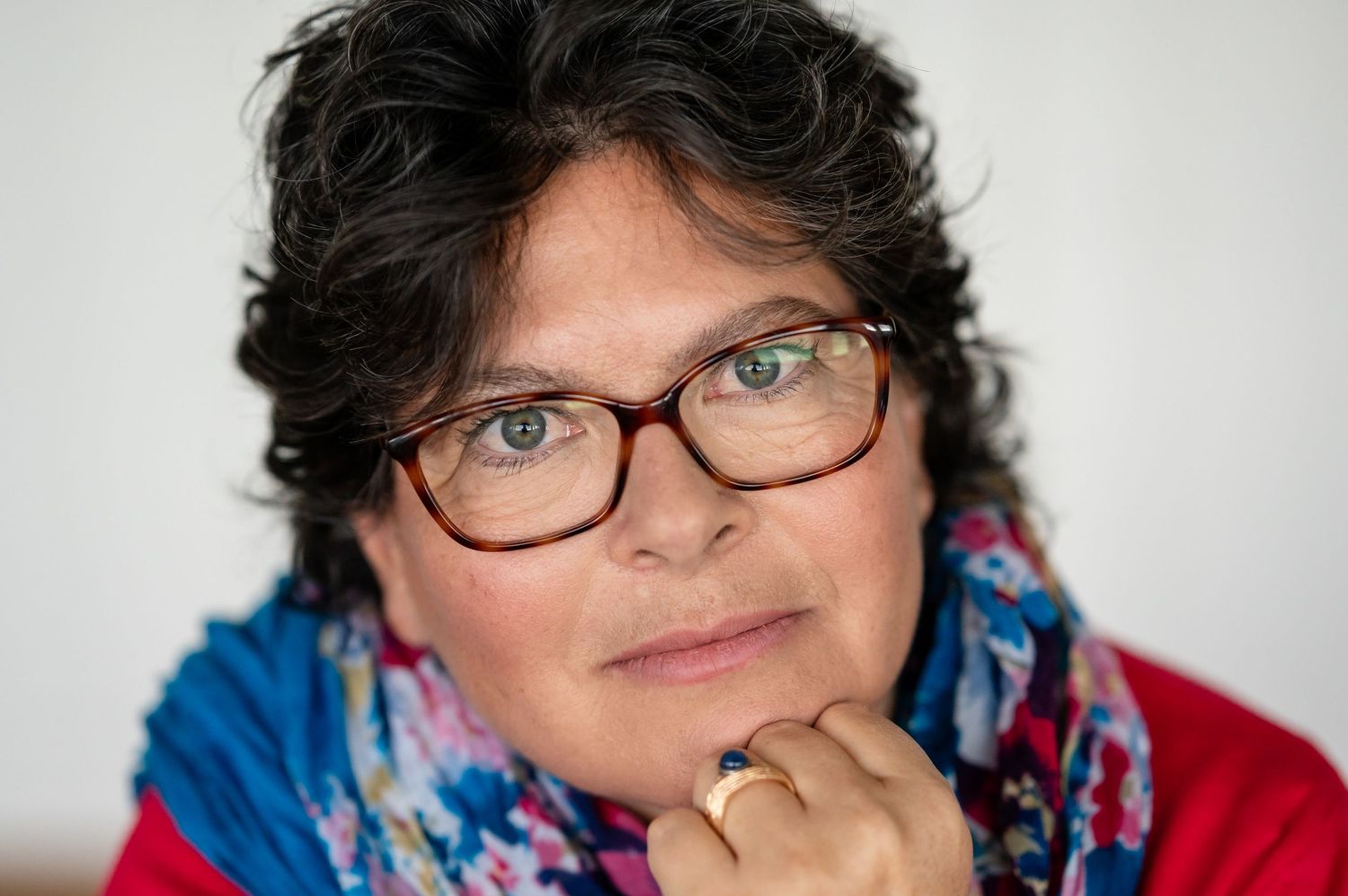 Porträt einer Frau mit Brille und lockigem, dunklem Haar, die in die Kamera blickt. Sie trägt einen bunten Schal und ein rotes Oberteil