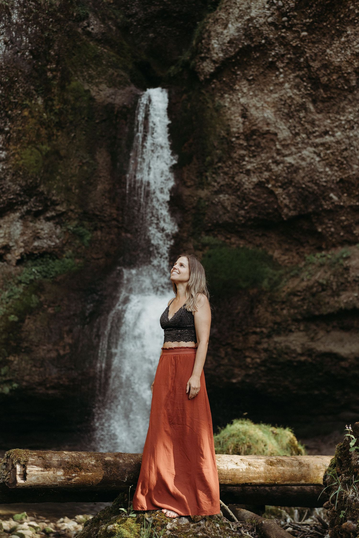 A waterfall cascades down rocky cliffs in a scenic forest setting with a rust-colored skirt in the foreground.
