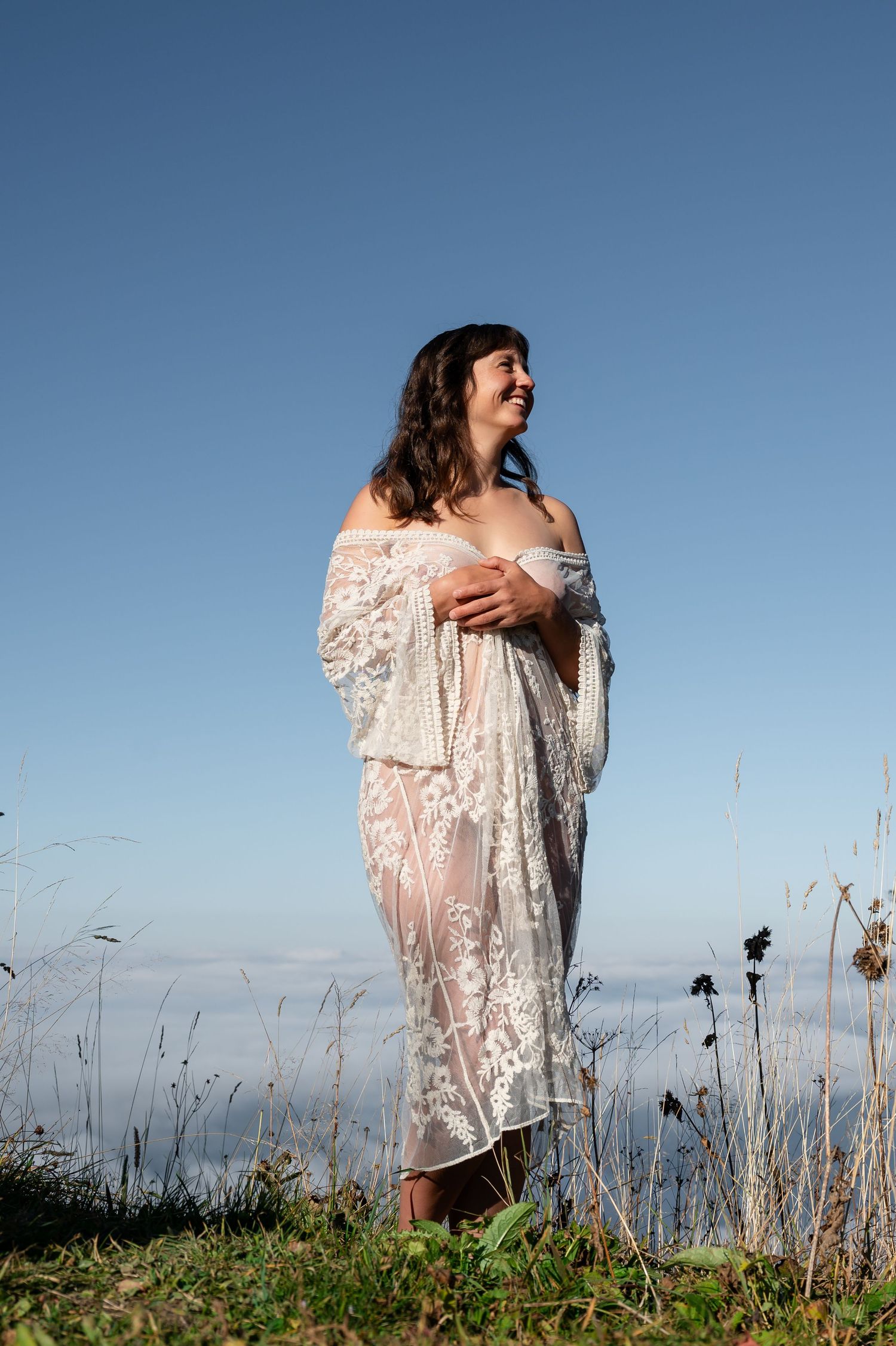 Frau in transparentem Spitzenkleid steht barfuß auf einer Wiese über den Wolken, umgeben von Himmel und Natur – Boudoir im Berner Oberland.