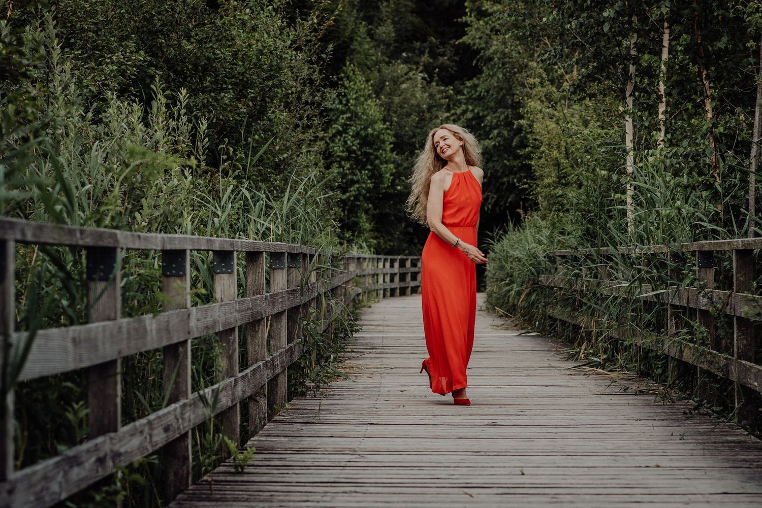 Frau im roten Kleid spaziert barfuß auf Holzsteg durch grüne Natur – ausdrucksstarkes Portrait mit Kontrast und Leichtigkeit.