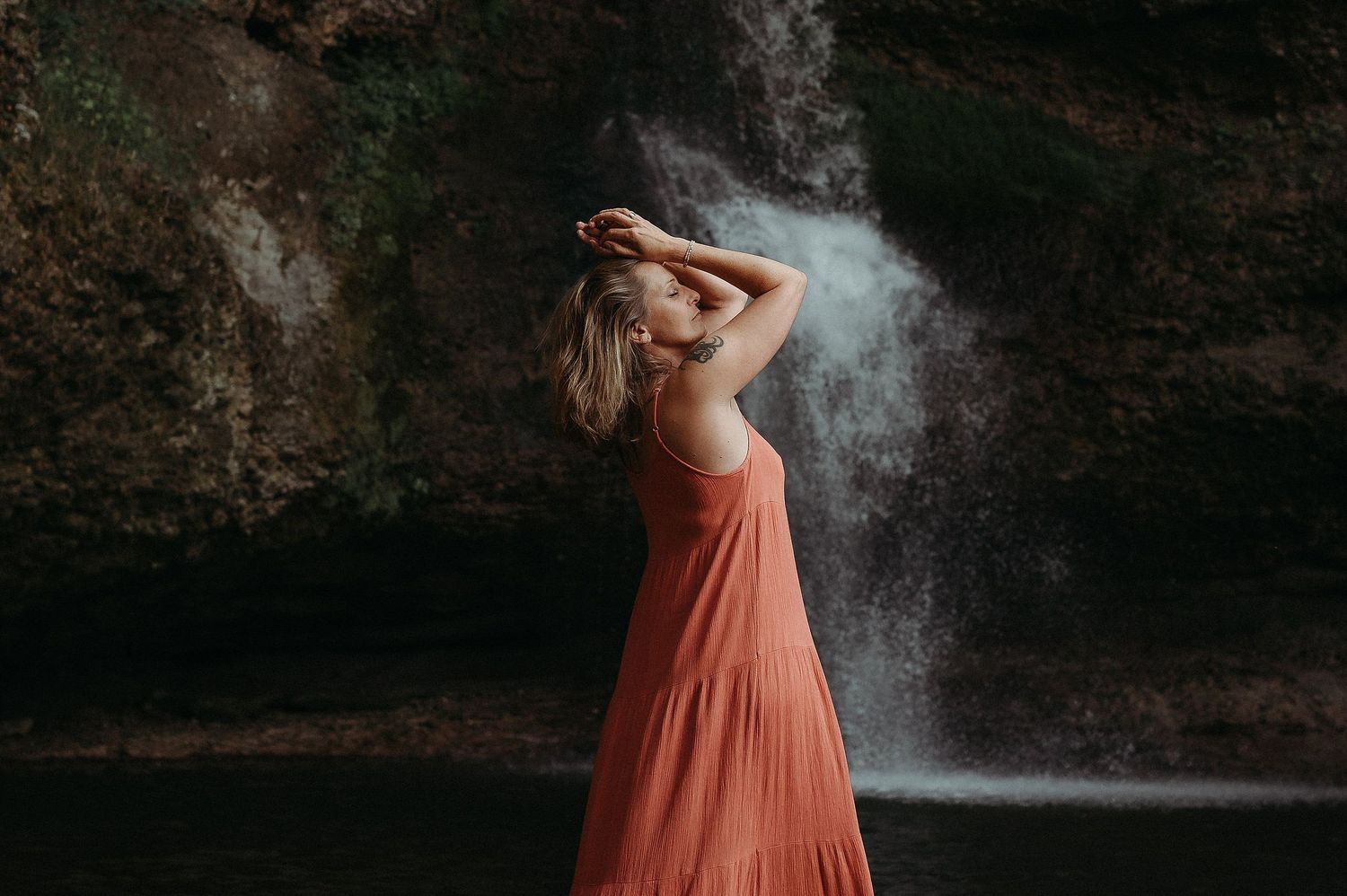 A figure in a flowing coral dress poses gracefully near a misty waterfall in a dramatic outdoor setting.
