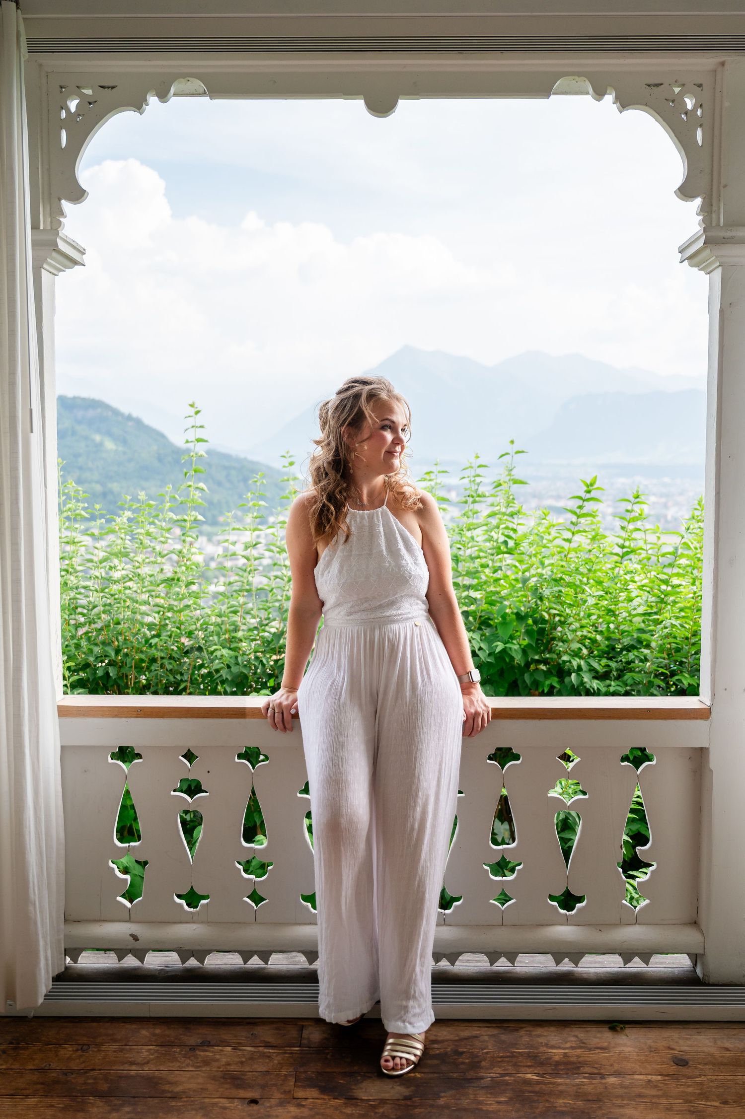 Frau im weissen Jumpsuit steht auf Balkon mit Blick auf Berge und Thunersee. Sommerliches Portraitshooting Nähe Thun, Bern.