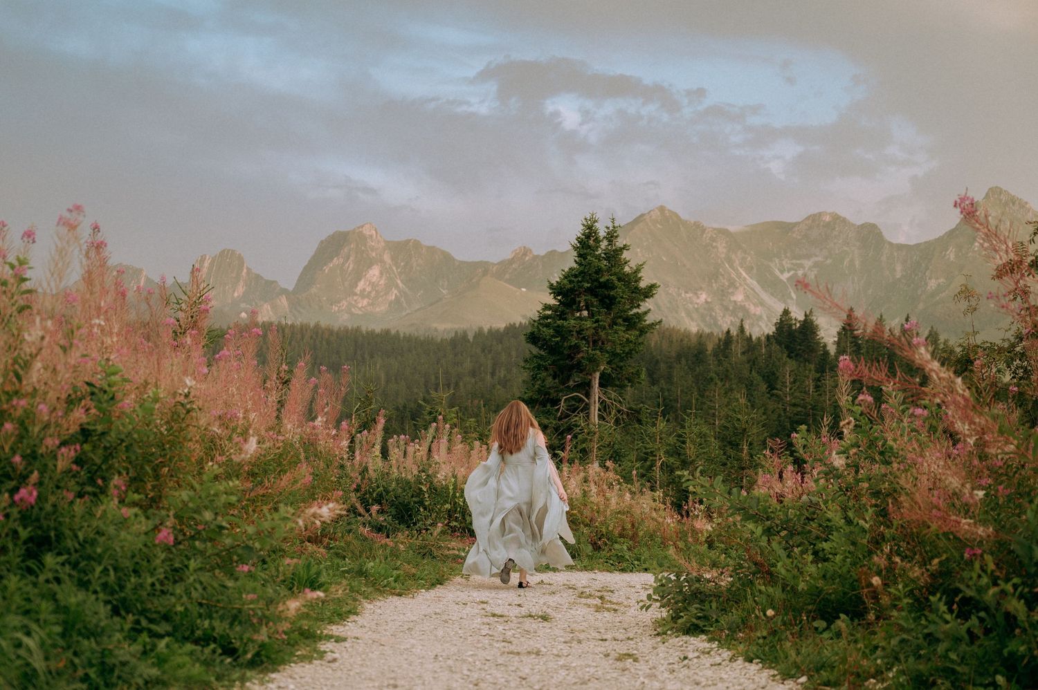 Frau im türkisen Kleid rennt auf einem Bergweg im Gantrisch, umgeben von Blumen, mit Blick auf ein dramatisches Alpenpanorama.
