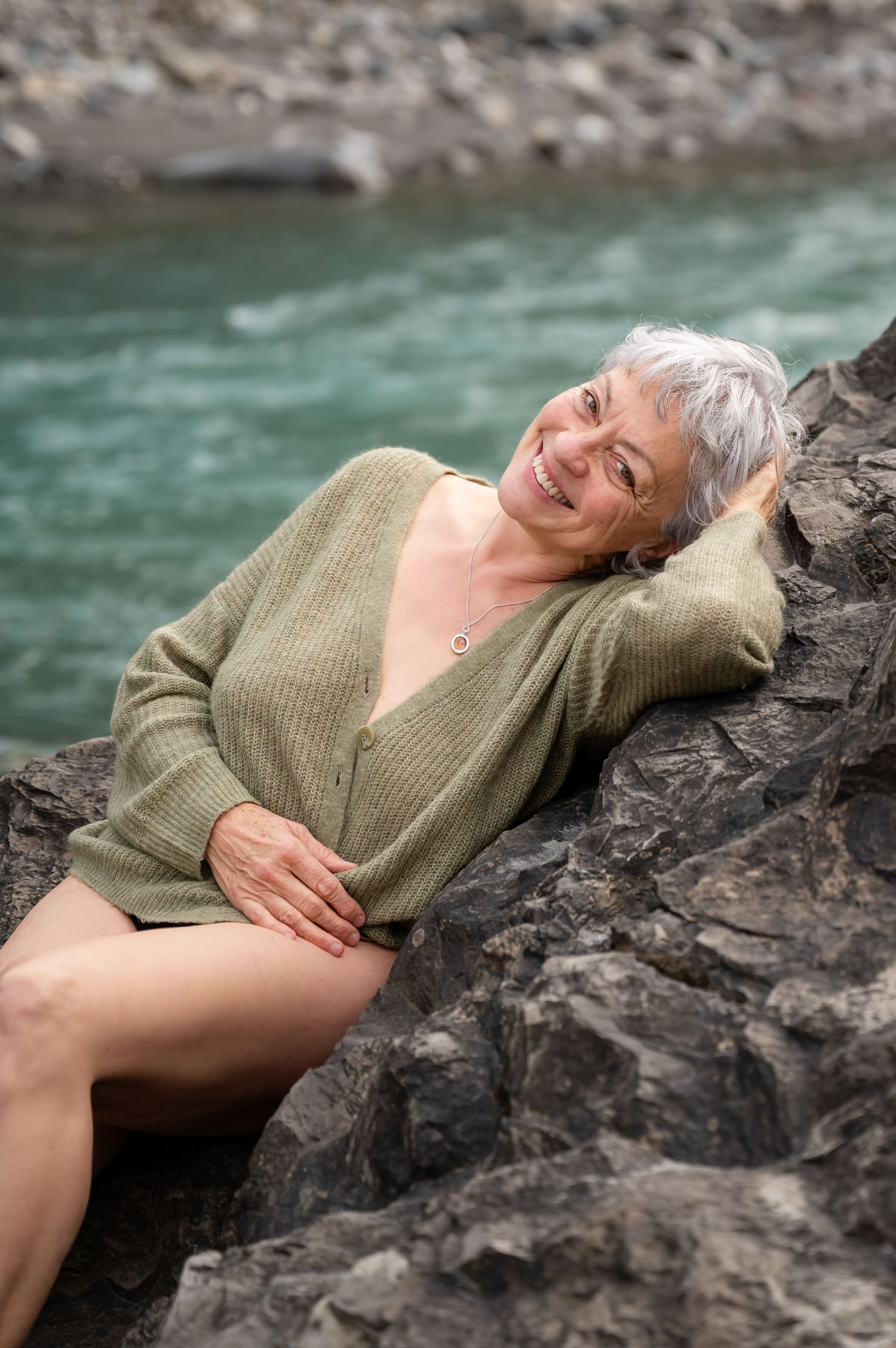 Ältere Frau beim Boudoir-Shooting am Fluss, entspannt und sinnlich in einem grünen Pullover auf Felsen liegend, selbstbewusst lächelnd.