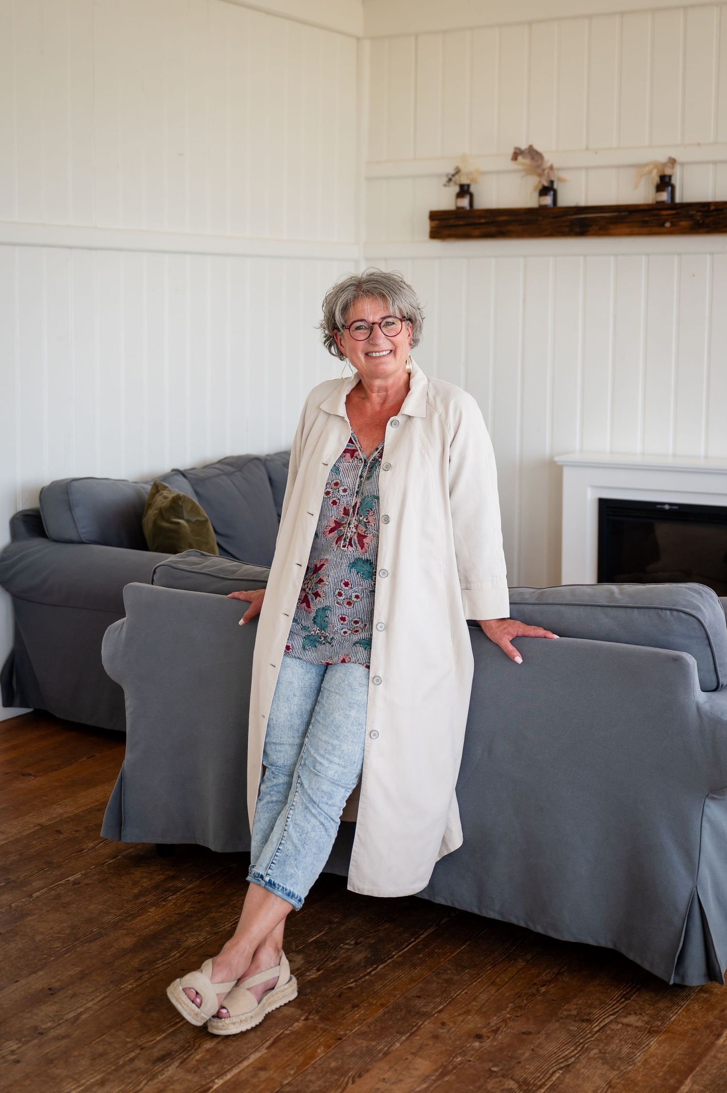 Ältere Frau mit grauen Haaren und Brille steht entspannt im Wohnzimmer, trägt einen langen Mantel und lächelt – Selbstliebe-Shooting