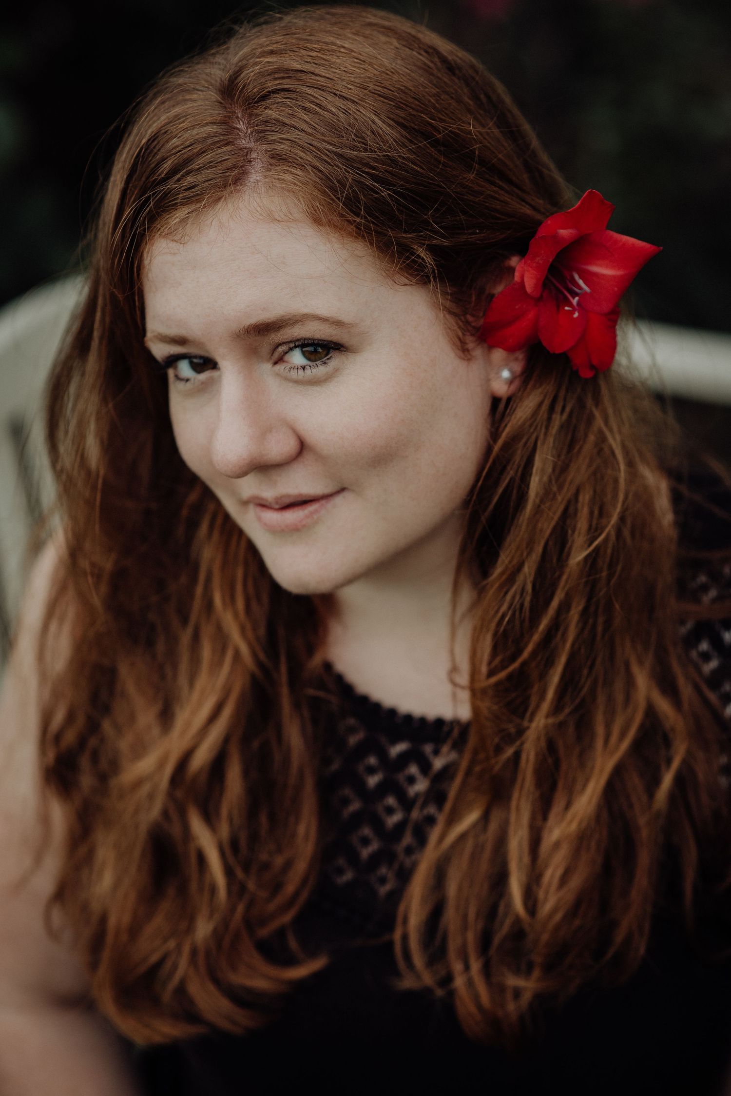 Frau mit langen roten Haaren und roter Blume im Haar, trägt ein schwarzes Top, natürlicher Portraitstil, aufgenommen in Thun.