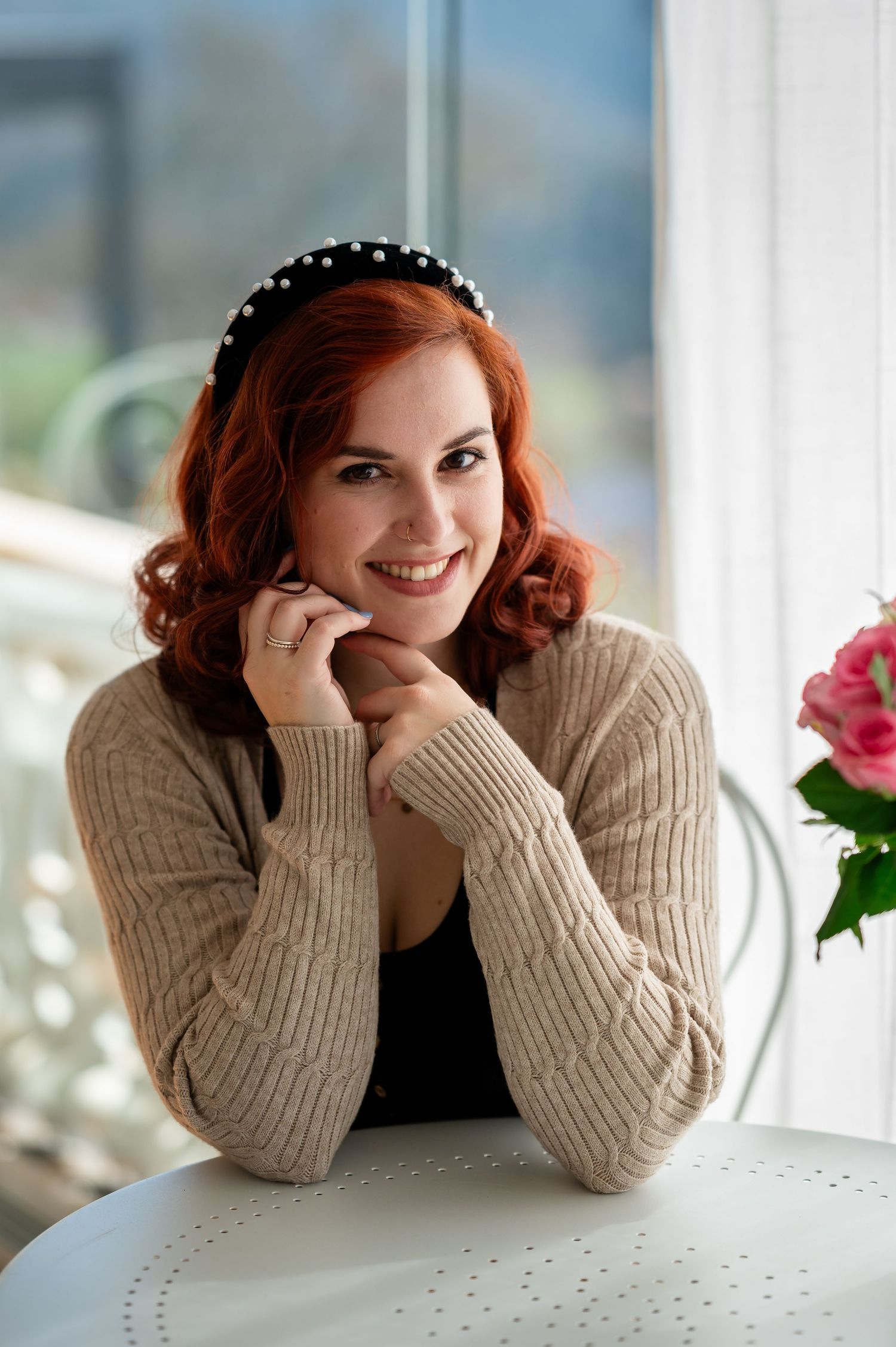 Junge Frau mit roten Haaren und Perlen-Haarband sitzt lächelnd an einem Tisch – Selbstliebe-Foto-Shooting in natürlichem Licht.