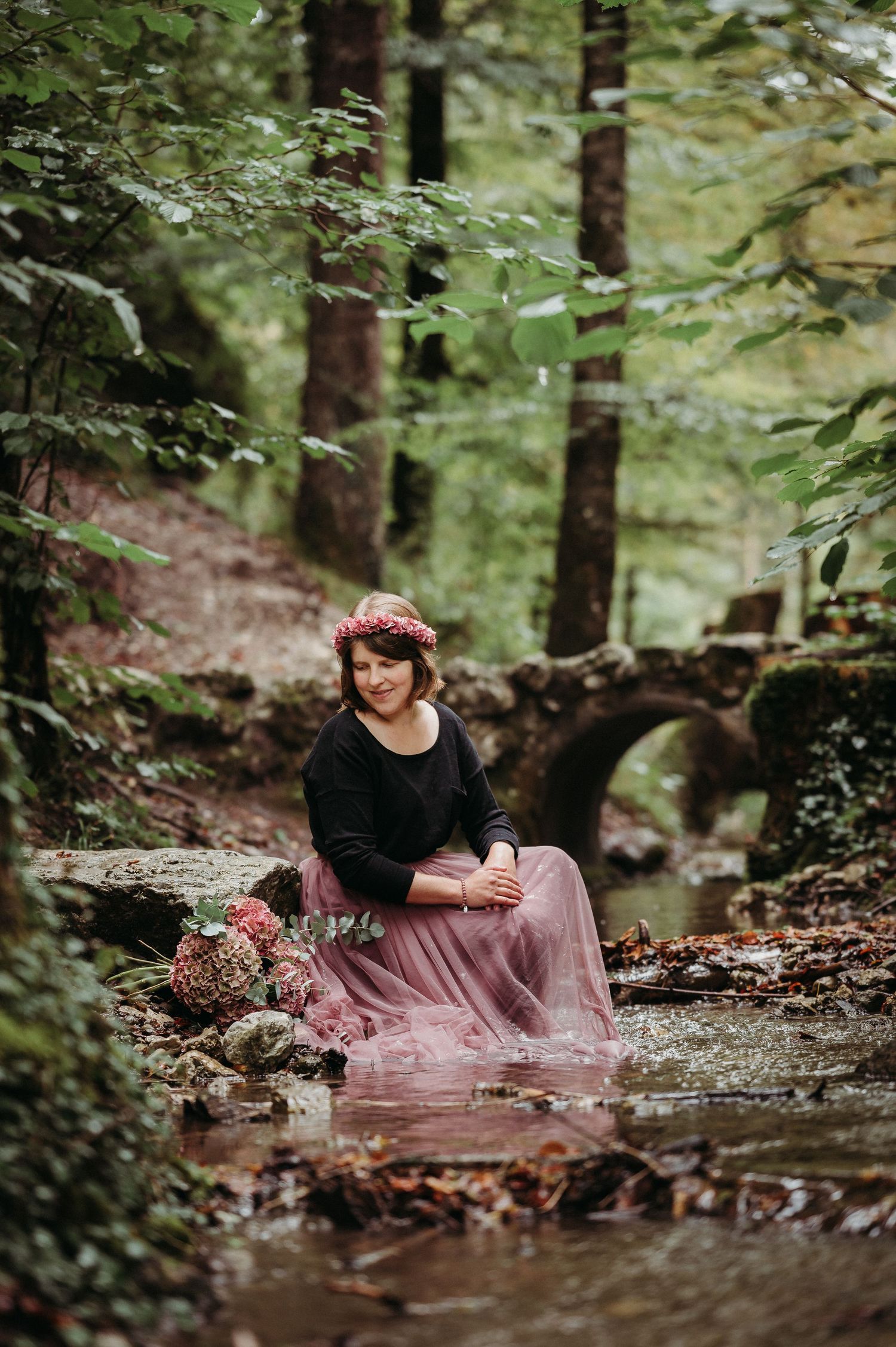 Frau mit Blumenkranz und Tüllrock sitzt am Bach im Strättligen Wald – märchenhaftes Portrait-Shooting in natürlicher Umgebung.