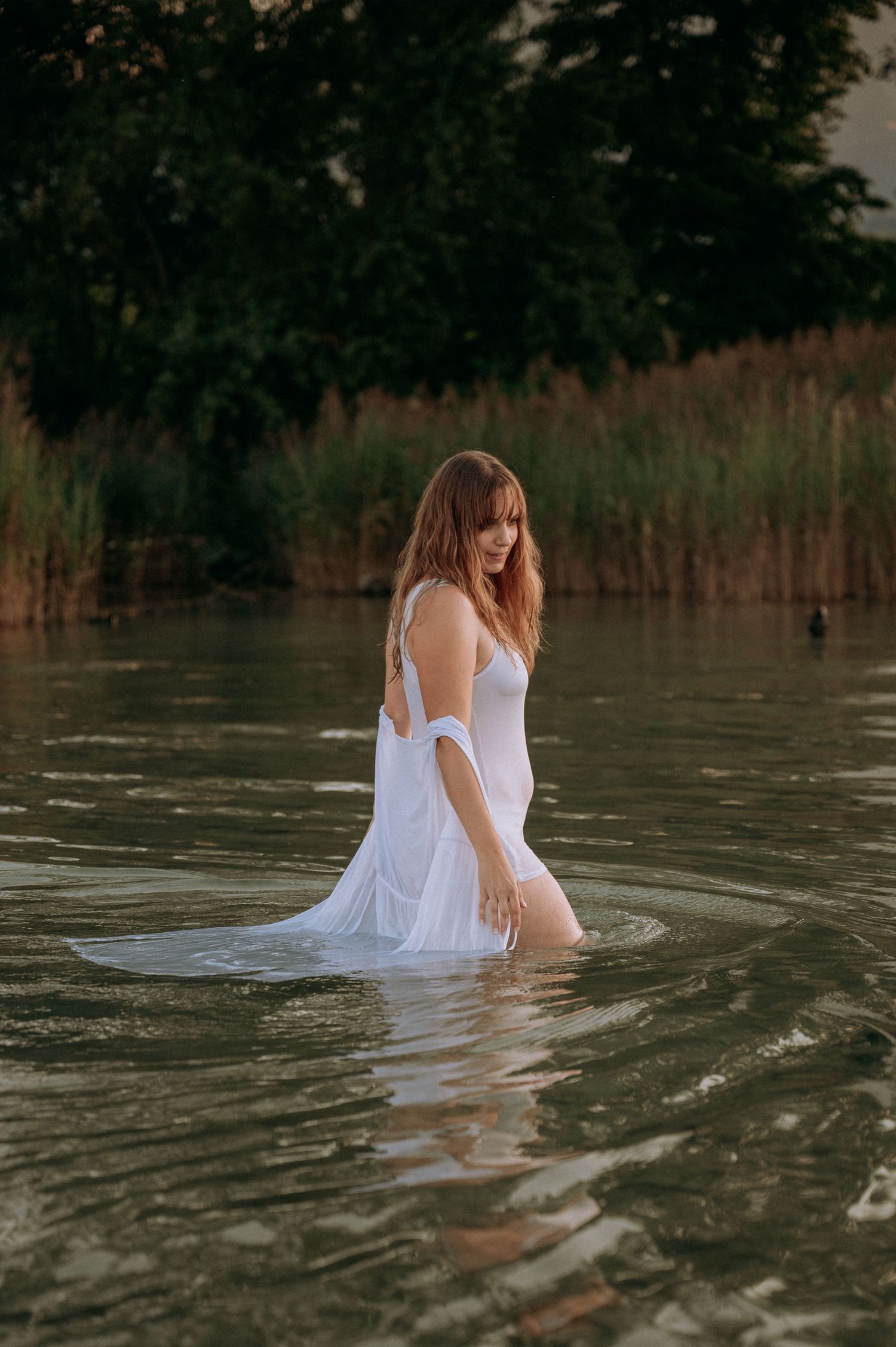 Boudoir-Shooting im See bei Sonnenuntergang: Frau in nassem, weißen Kleid watet sinnlich durchs Wasser – natürlich, ruhig, feminin.