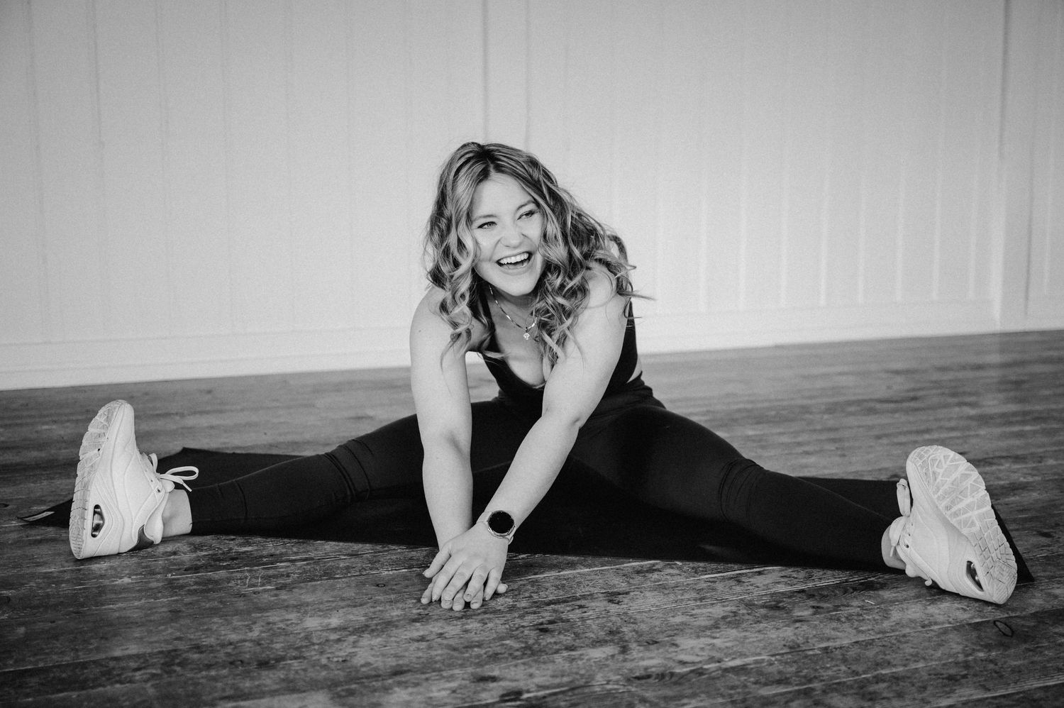 A person in black athletic wear does a full split stretch on a wooden floor in a black and white photograph.