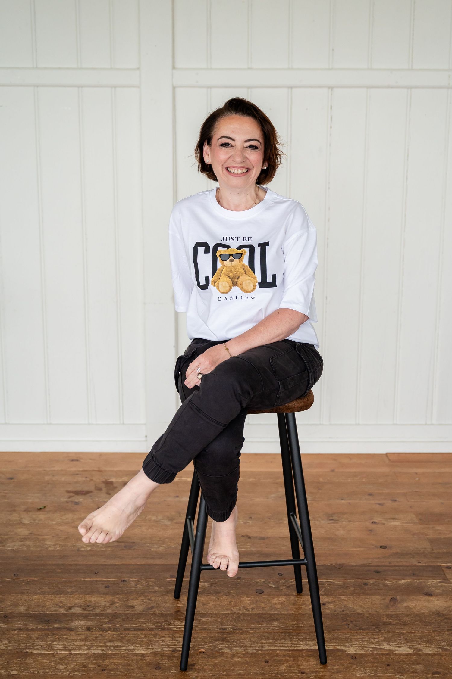 Lachende Frau beim Selbstliebe-Shooting in Thun, barfuß auf einem Hocker, weißes Shirt mit Aufdruck, helle Holzlocation.
