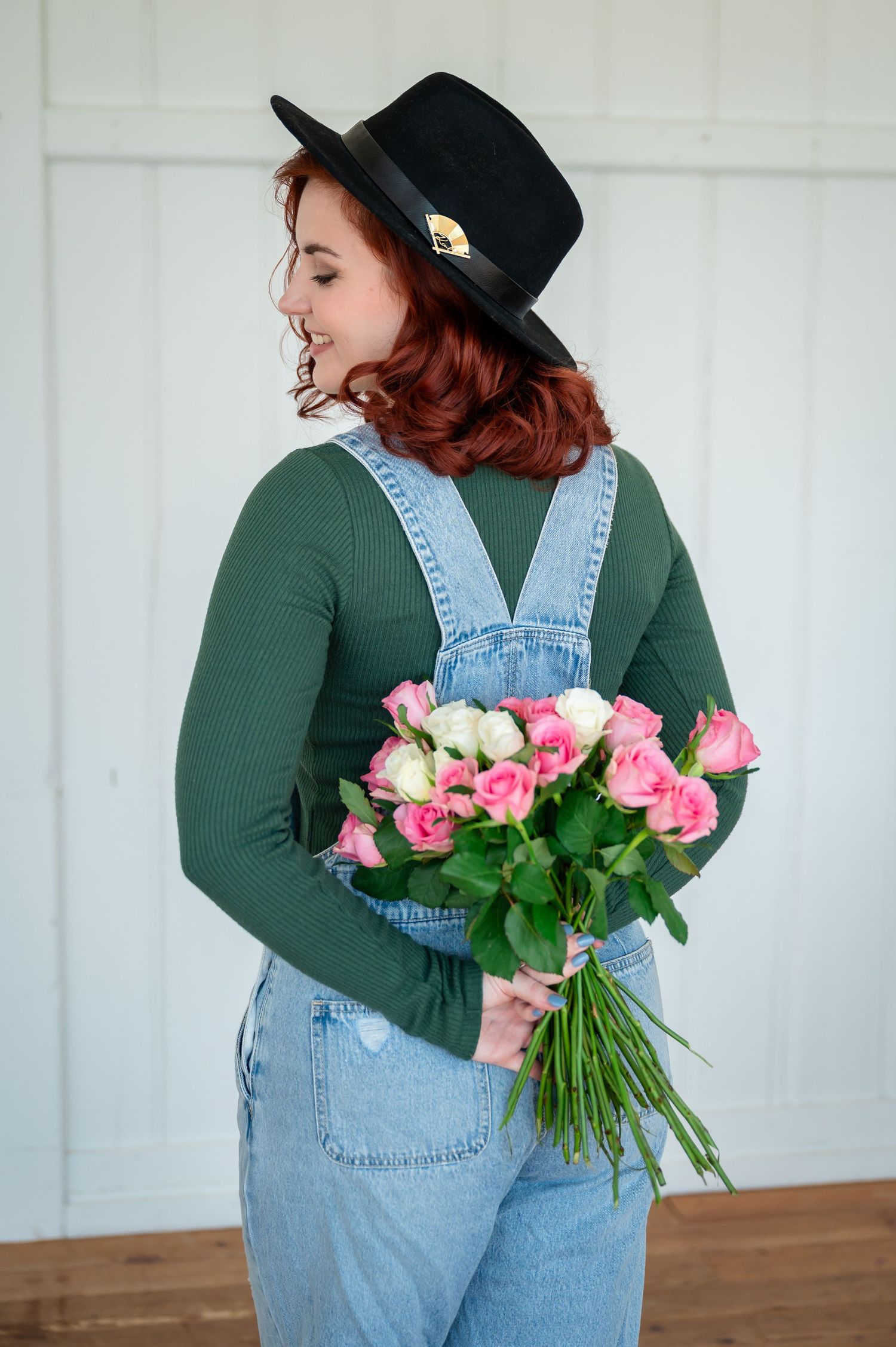 Junge Frau mit schwarzem Hut, grünem Oberteil und Latzhose hält einen Strauss rosa Rosen hinter dem Rücken –Selbstliebe-Shooting