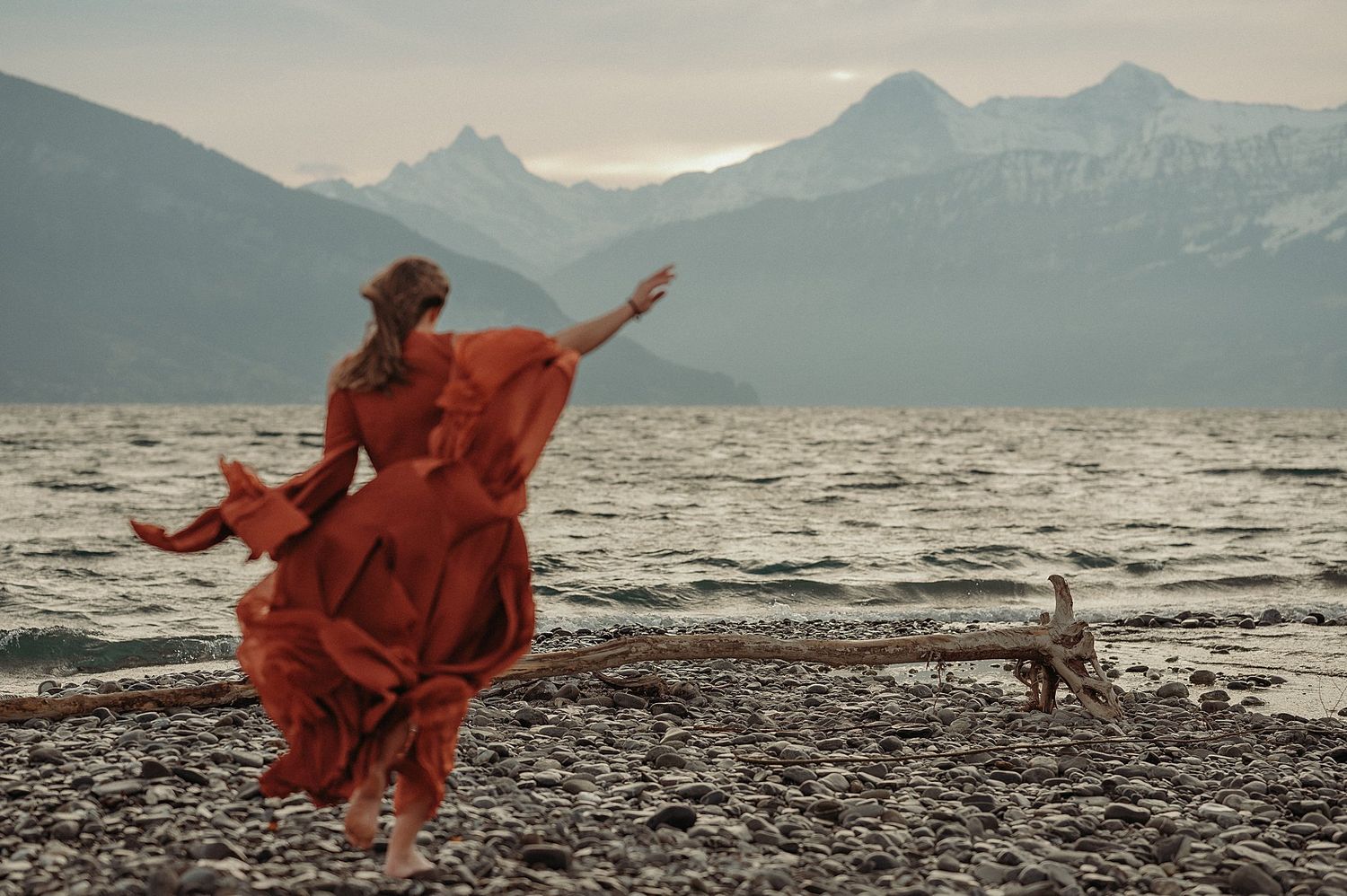 A flowing red dress billows in the wind on a rocky beach with snow-capped mountains in the background at dusk.
