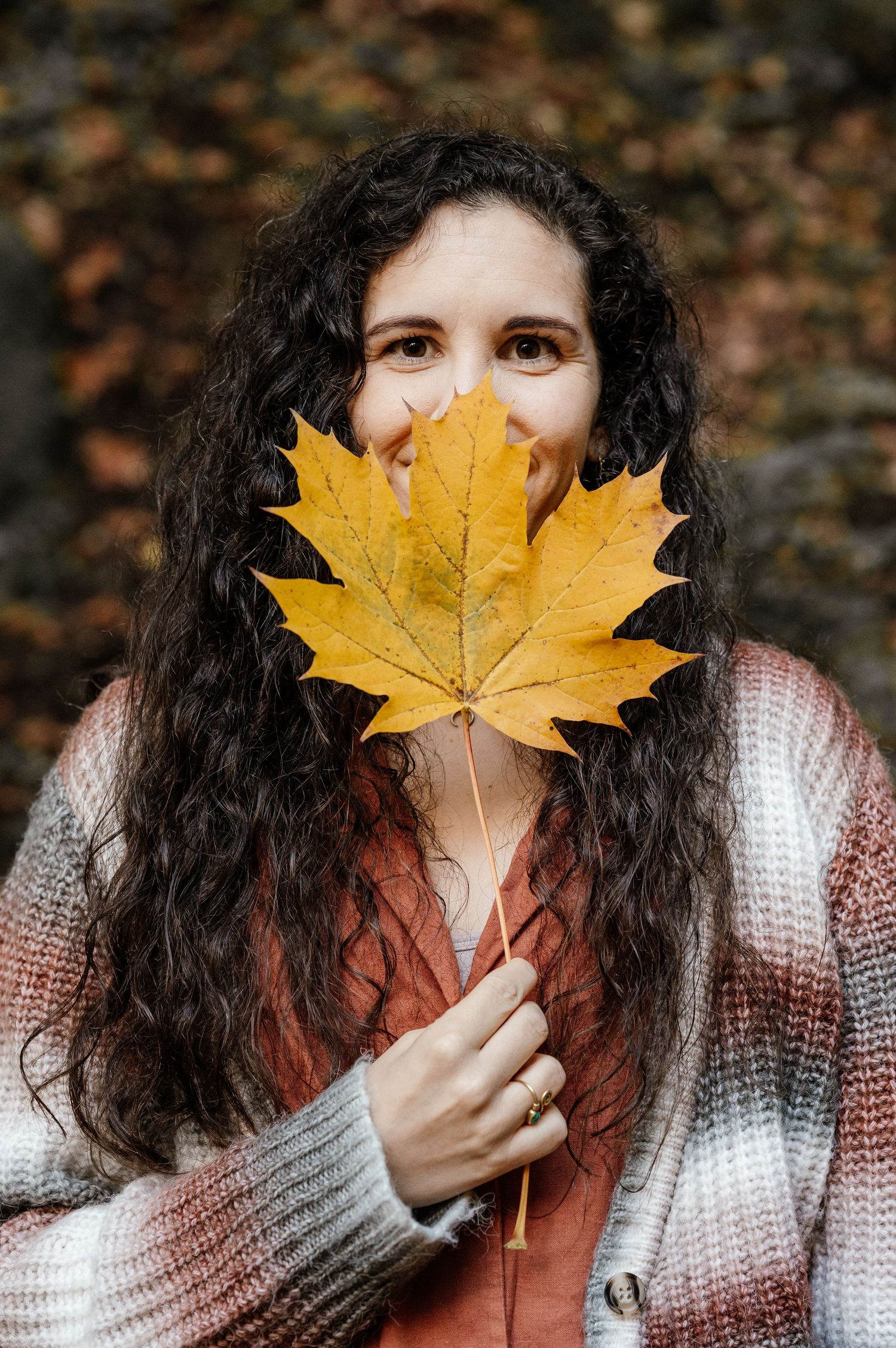 Frau hält ein großes Ahornblatt vor ihr Gesicht – herbstliches Portrait mit bunten Farben und verspielter, fröhlicher Stimmung im Wald.