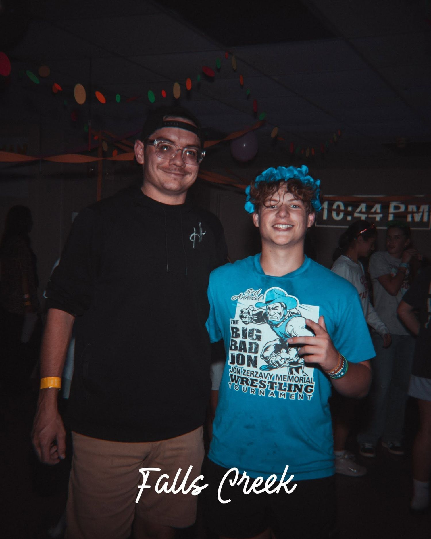 Two young men smiling at Falls Creek camp event at 10:44 PM, one wearing a teal Big Bad John wrestling shirt.