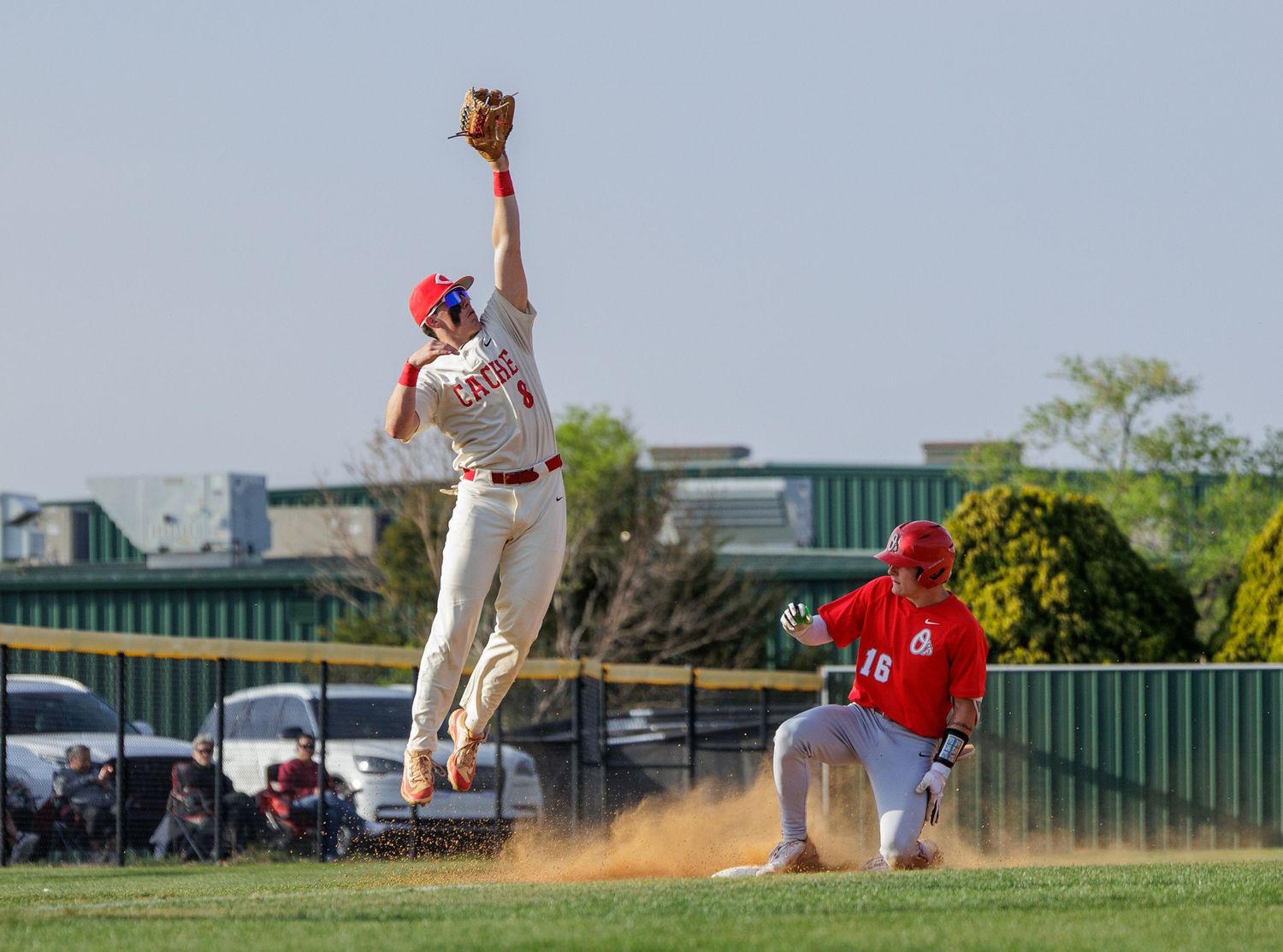 Baseball infielder leaps high to catch ball while runner slides into base in action shot.