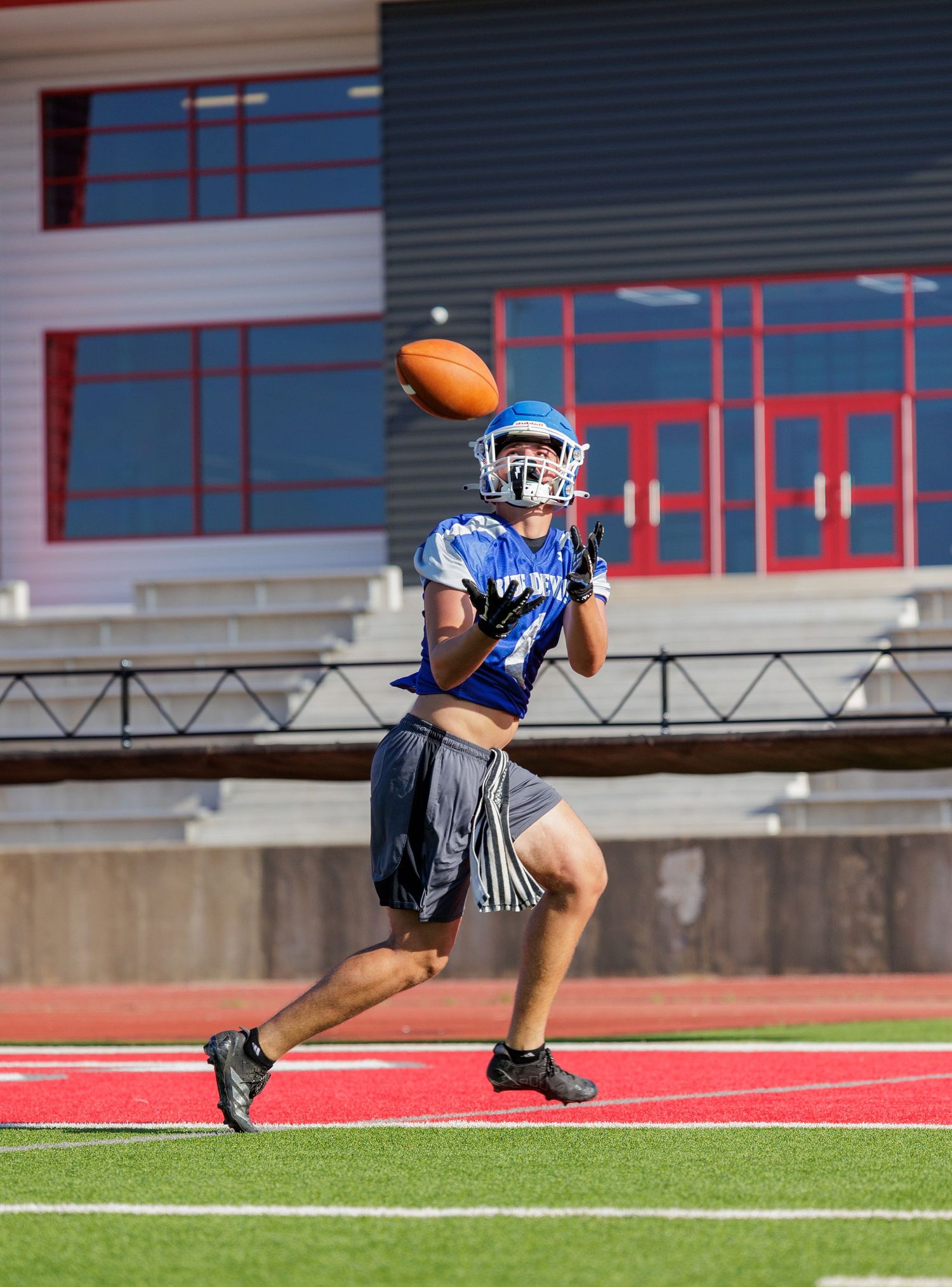 Football player in blue uniform reaches up to catch a pass during an outdoor practice at a stadium.