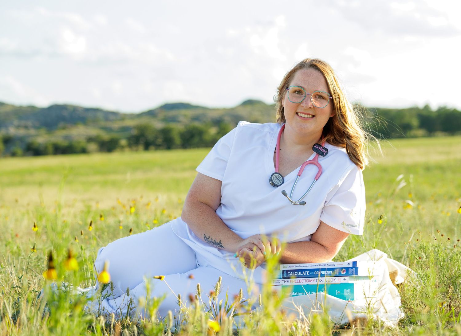Smiling nurse in white scrubs sits in a wildflower meadow holding medical books with hills in background.