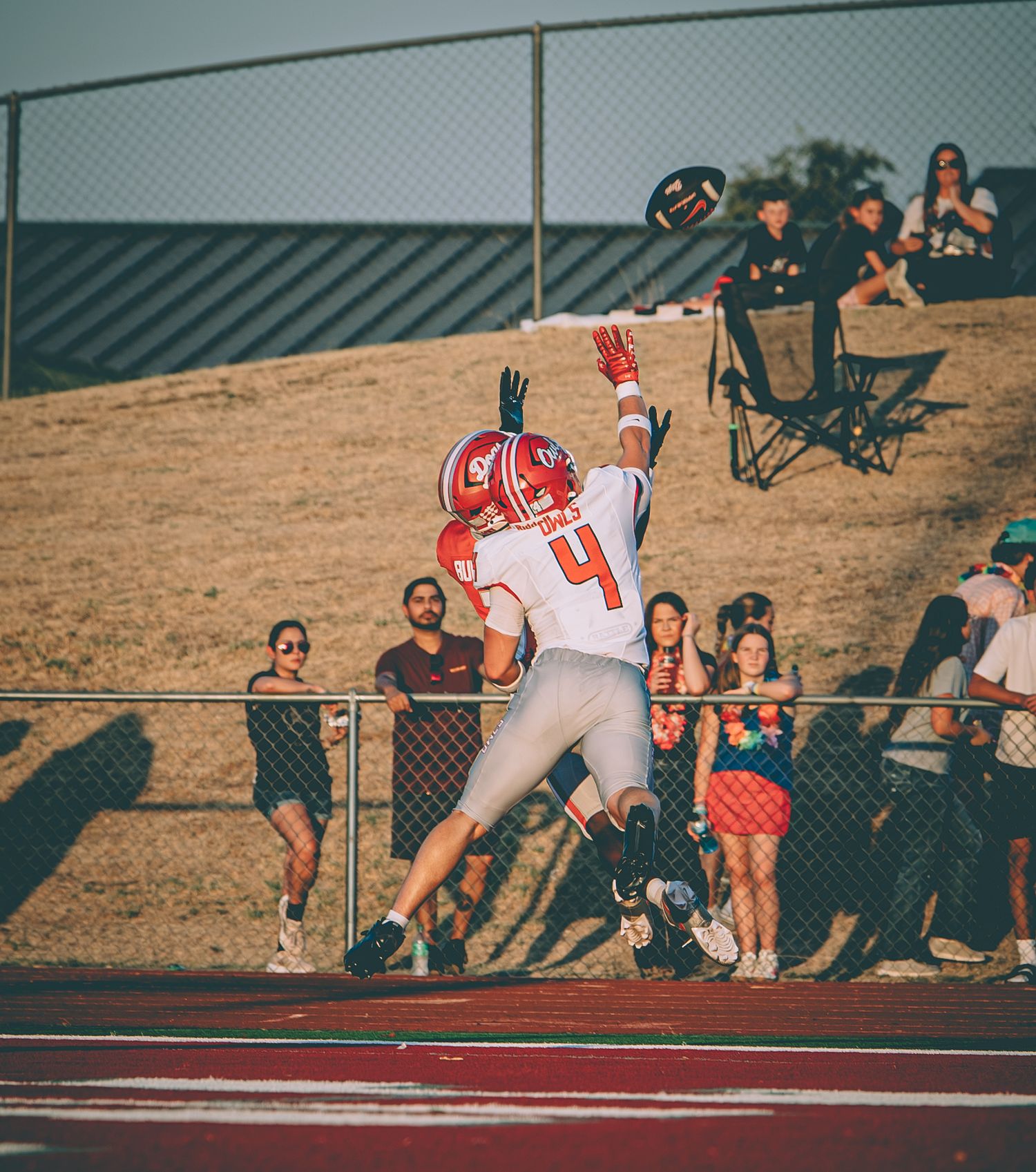 Football player #4 in orange jersey leaps to catch a pass during a game with fans watching from the sidelines.