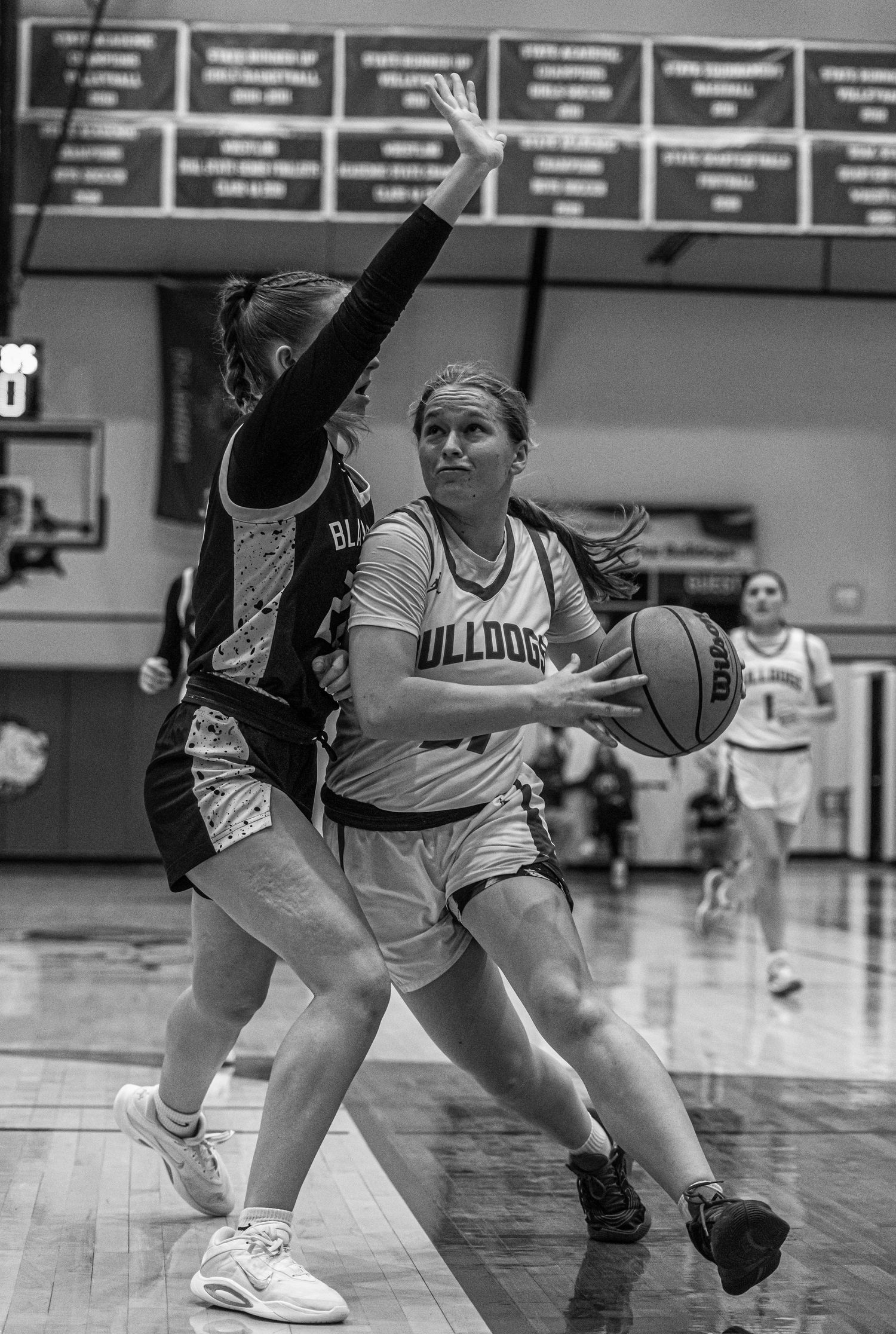 Female Bulldogs basketball player drives to basket against defender in black and white gym photo.