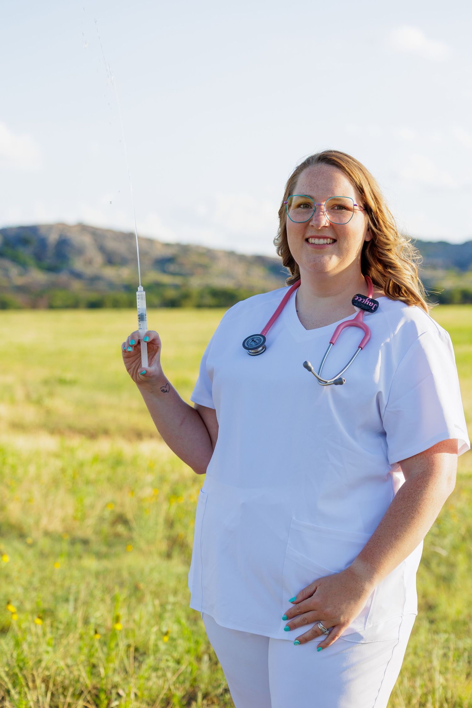 Female nurse in white scrubs holding a syringe and stethoscope outdoors in a green meadow.
