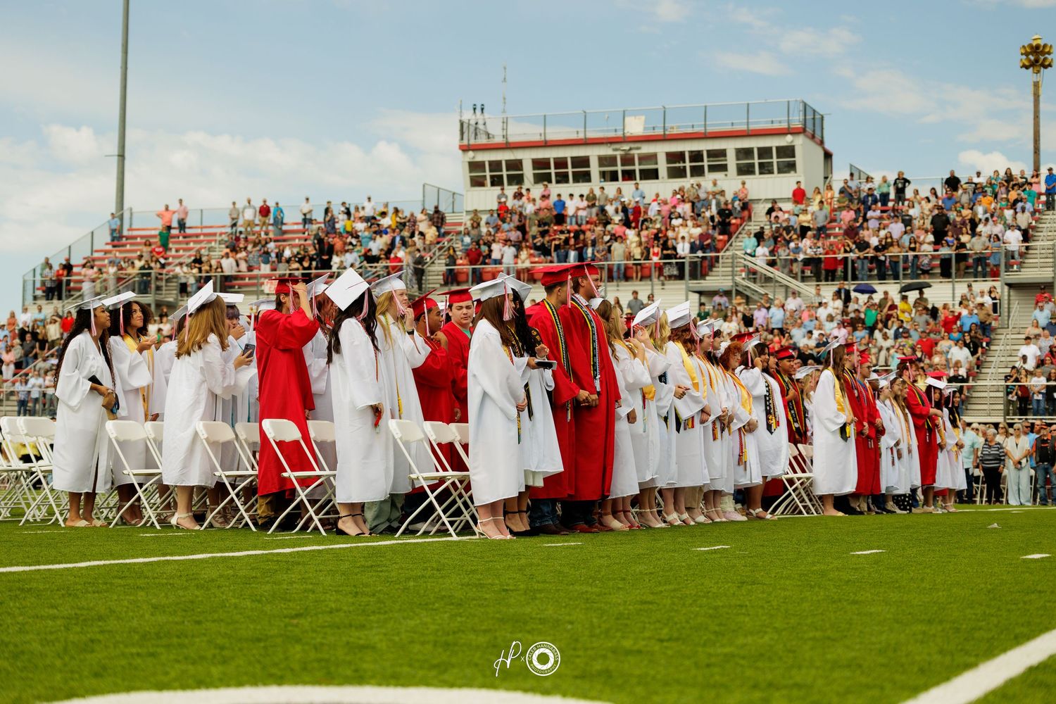 Graduation ceremony with students in red and white caps and gowns standing on a football field stadium.