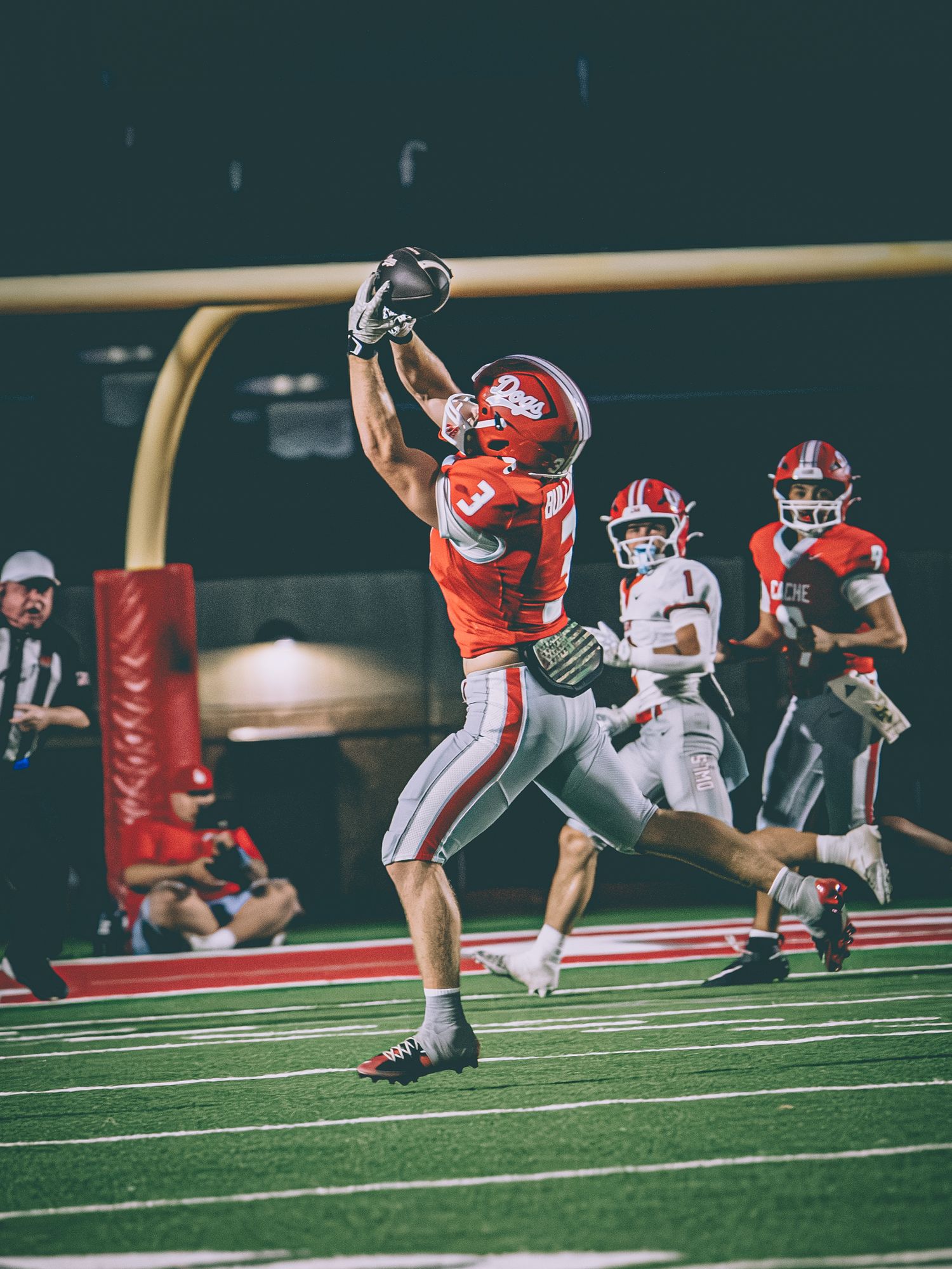 Football player in red jersey #3 makes a leaping catch during a night game on a red turf field.
