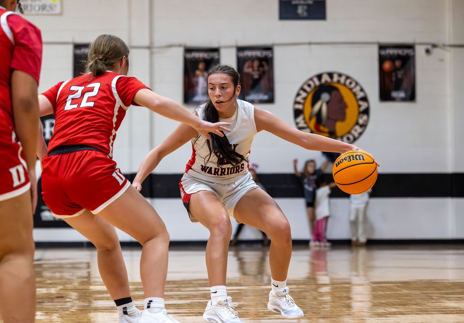 Female basketball player in white Warriors jersey dribbles ball while defended by opponent wearing number 22 red uniform.