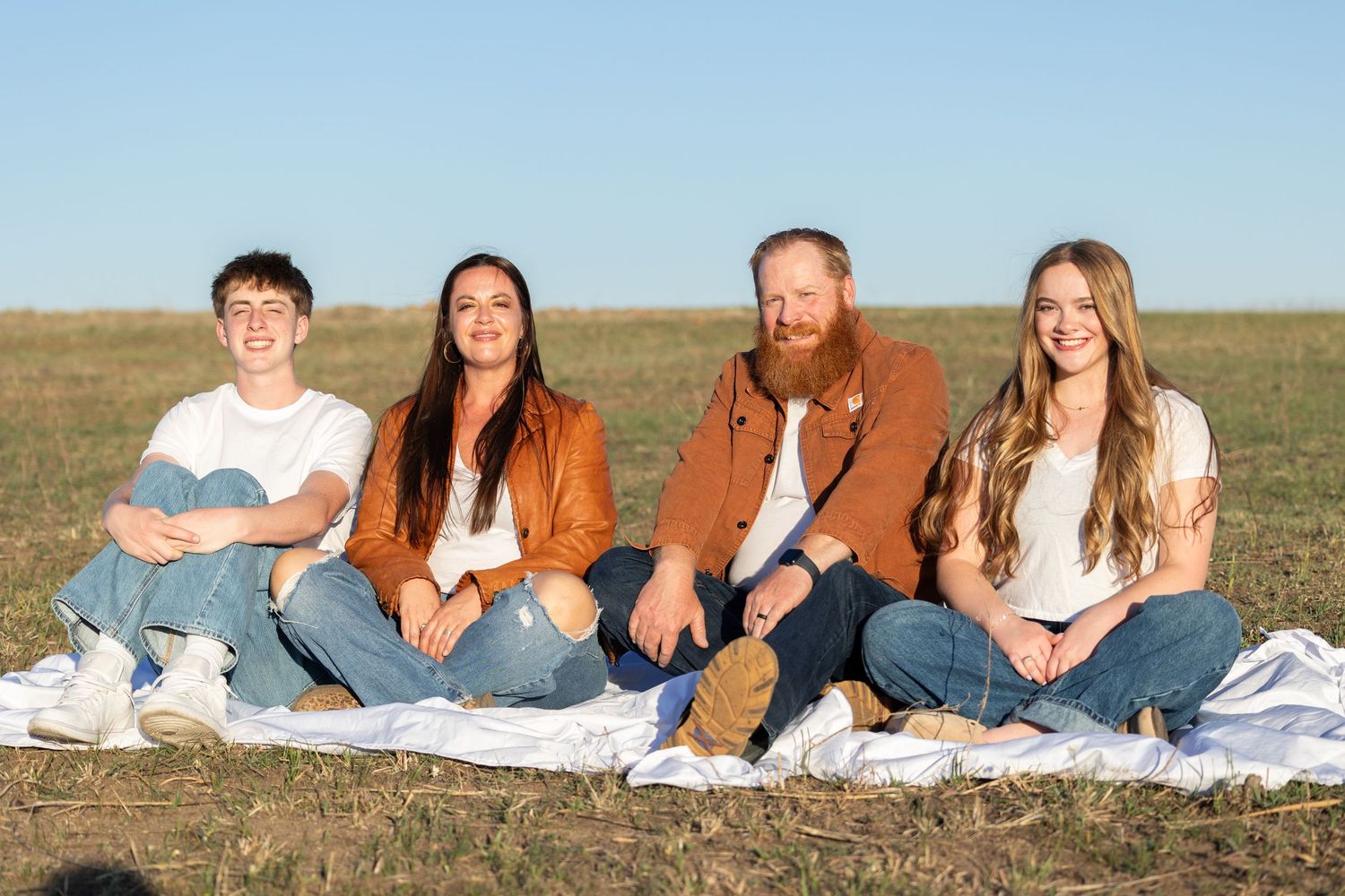 Happy family of four sits together on a white blanket in an open field under a clear blue sky.