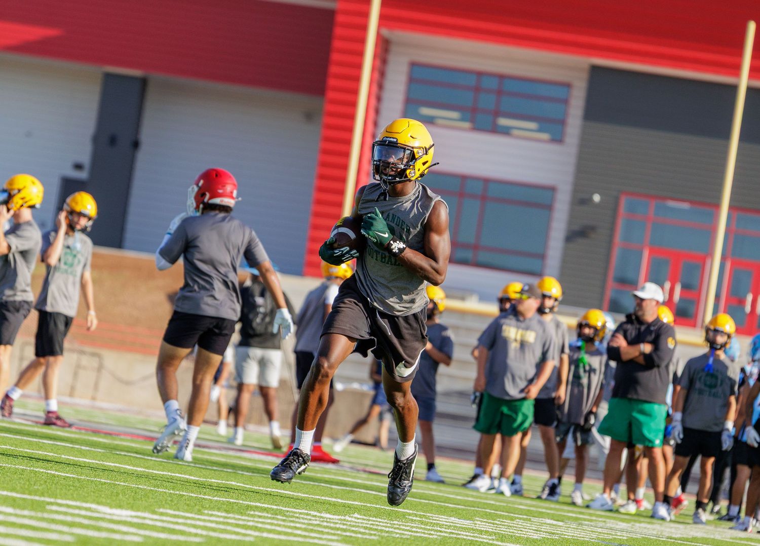 Football player in yellow helmet runs with the ball during a team practice session at a stadium.