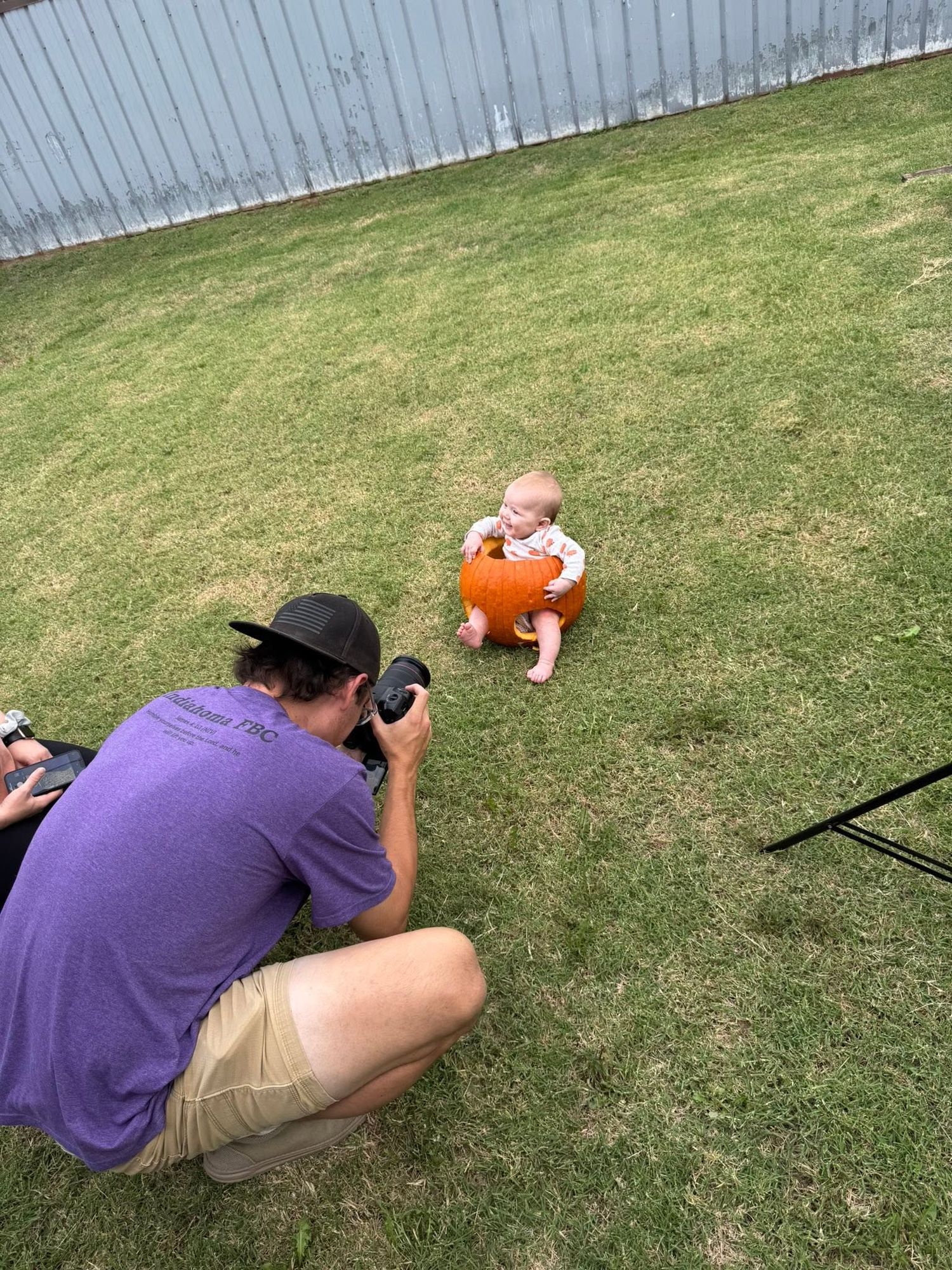 Photographer in purple shirt crouching to photograph a baby sitting in grass holding a pumpkin.