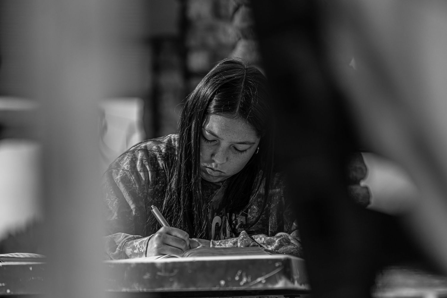 Black and white photo of young girl writing at desk in classroom setting.