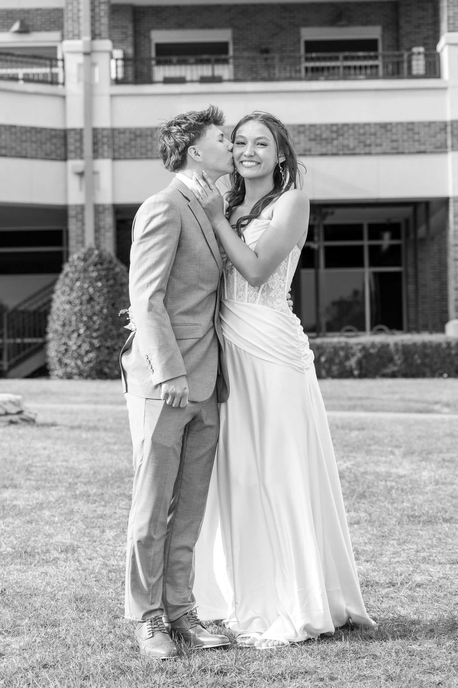 Black and white photo of a young couple in formal attire sharing a kiss outside a brick building.