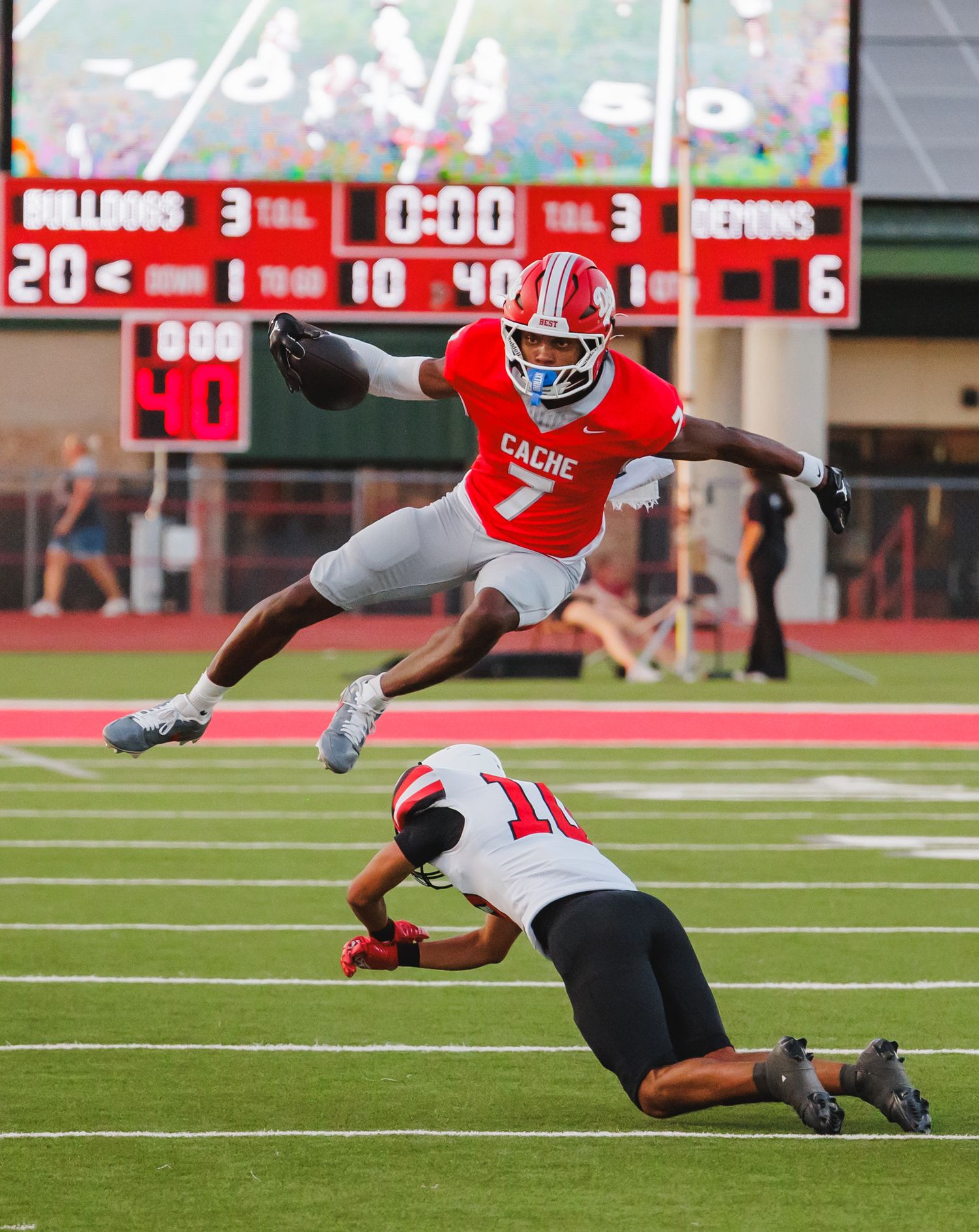 Cache Bulldogs football player #7 leaps over a sliding defender during a high school game with scoreboard visible.
