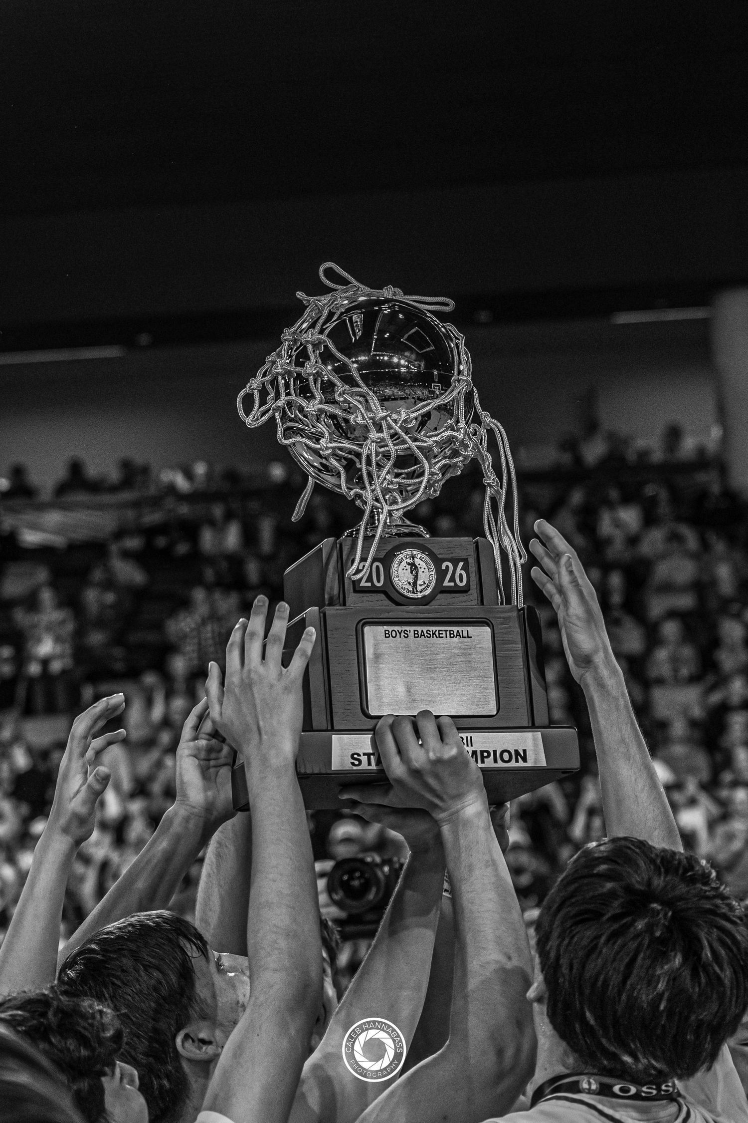 Players raise a boys basketball state champion trophy with net in black and white celebration photo.
