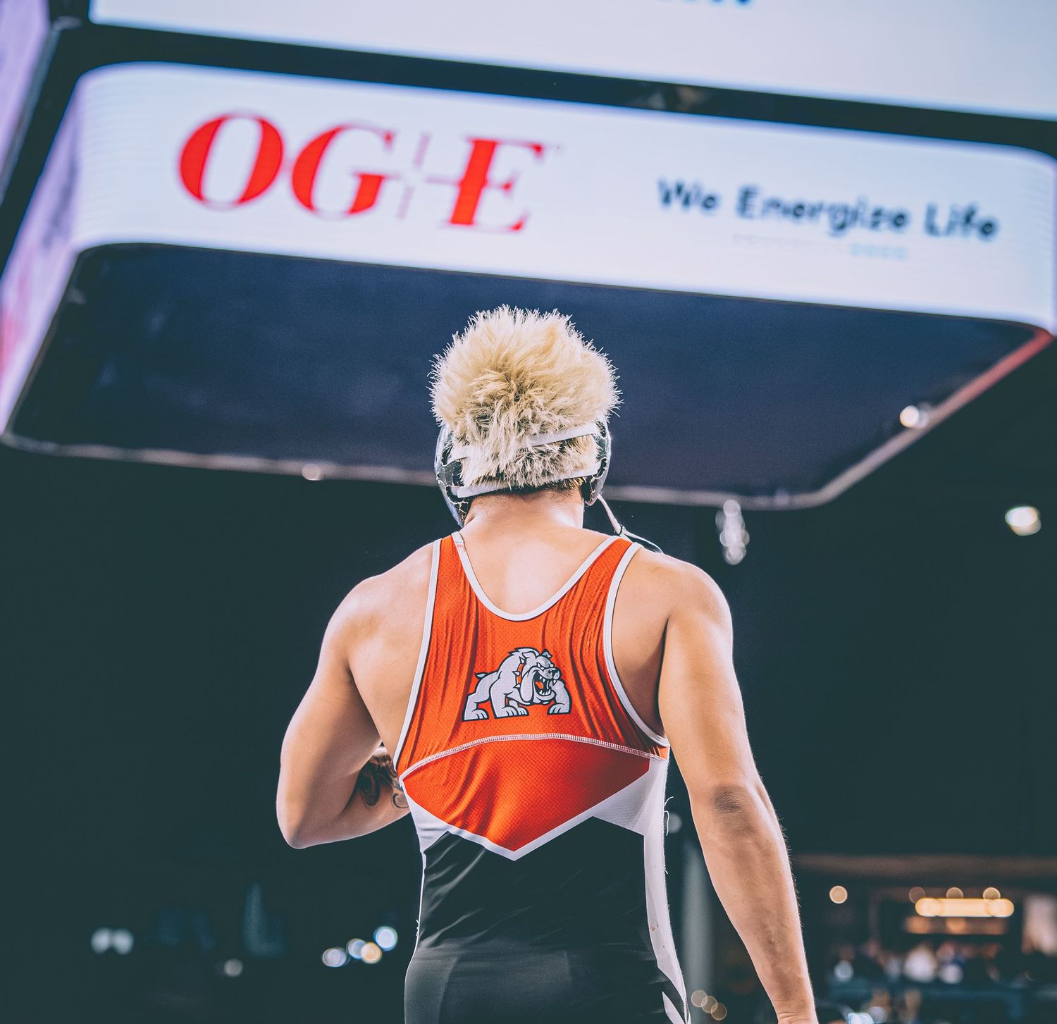 A wrestler in an orange singlet stands with back turned under an OGE Energy scoreboard at a wrestling competition.