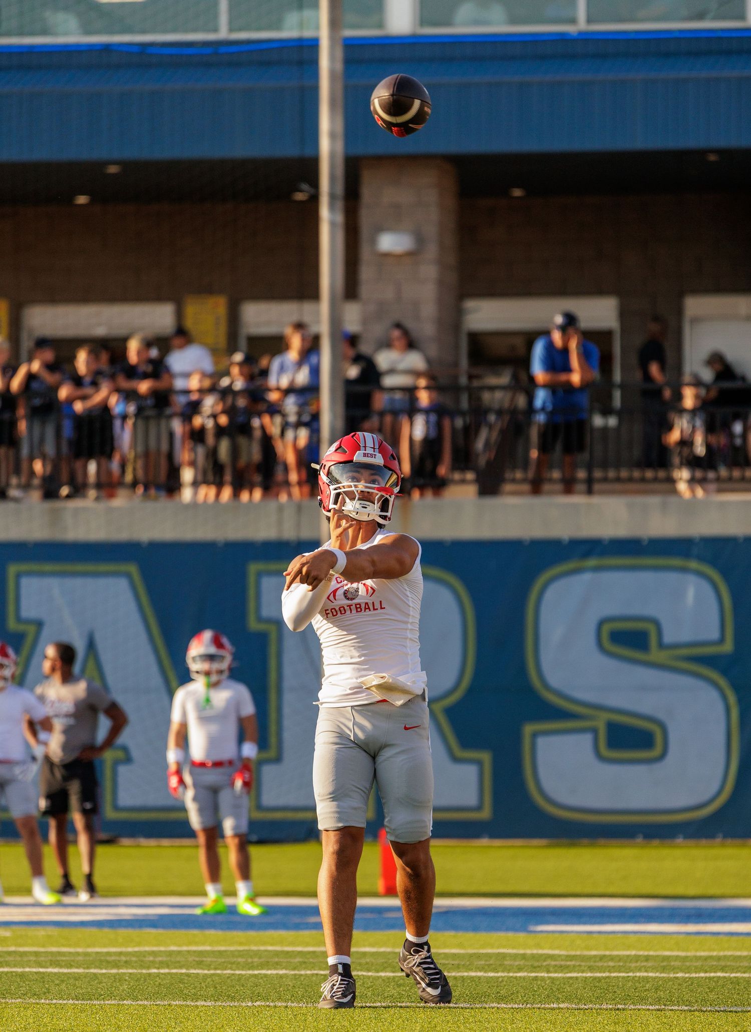 High school football player in white uniform throws a pass during a game at a blue stadium.