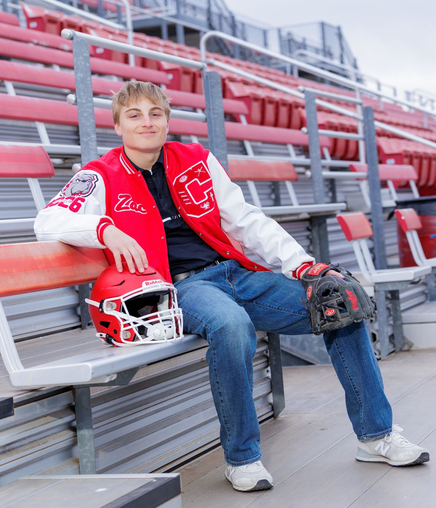 High school senior in red letterman jacket sitting in bleachers with football helmet and baseball glove.
