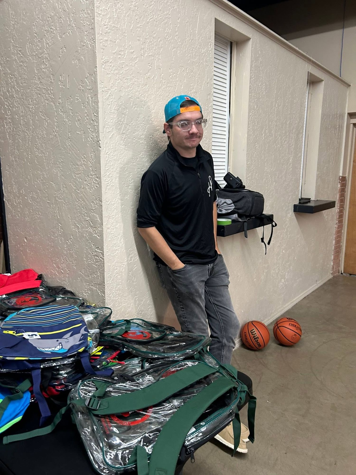 Young man with colorful hair and glasses leans against a wall near sports bags and basketballs indoors.
