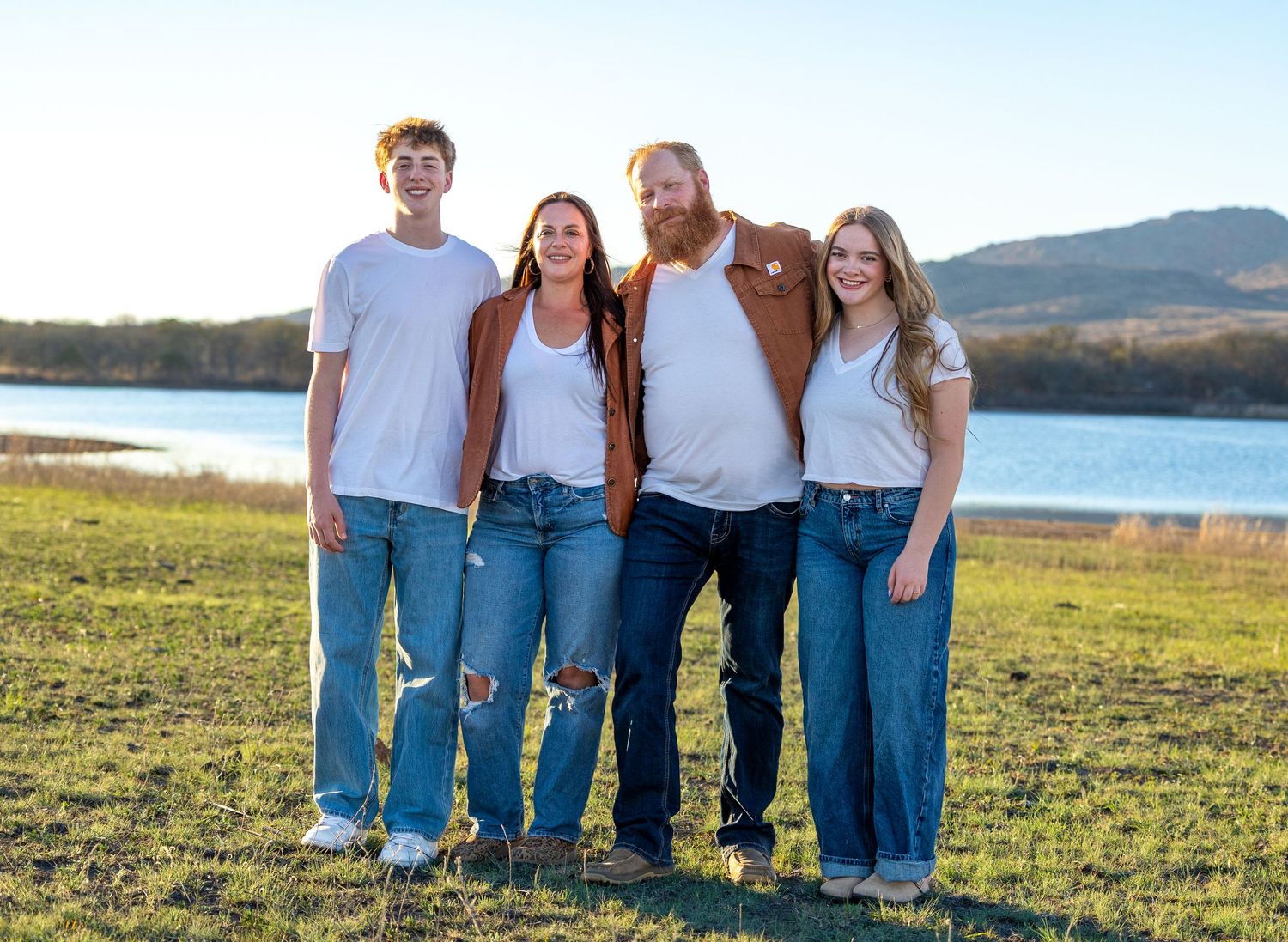 Family of four poses together smiling near a lake with mountains in the background at golden hour.