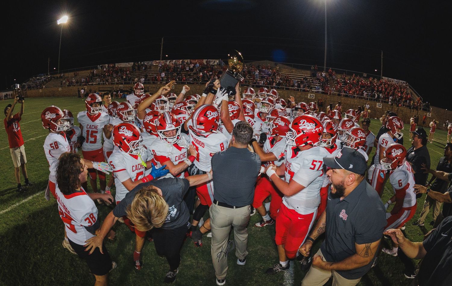 Red-uniformed football team celebrates victory with trophy raised under stadium lights at night.