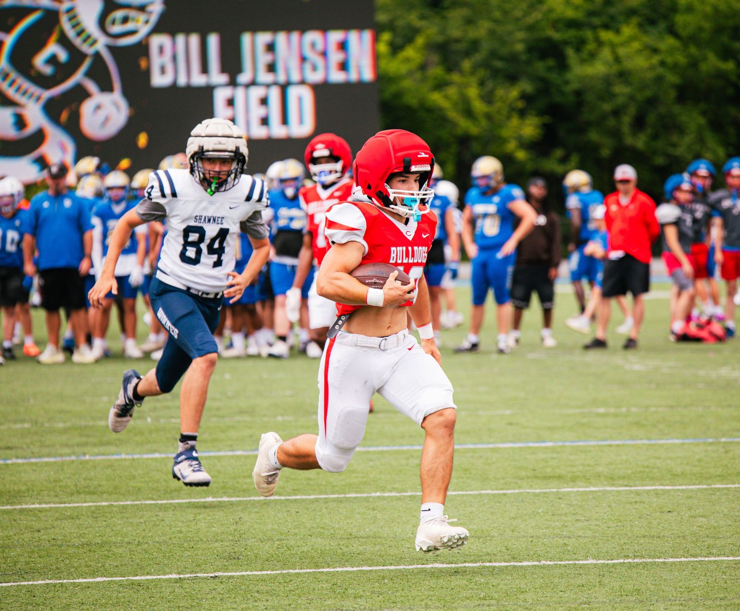 A football player in a red Bulldogs helmet carries the ball on Bill Jensen Field during a sunny outdoor game.