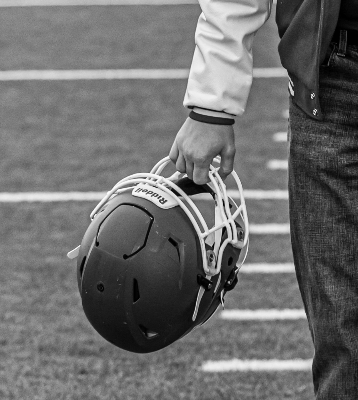 Black and white photo of a person holding a Riddell football helmet on a field sideline.