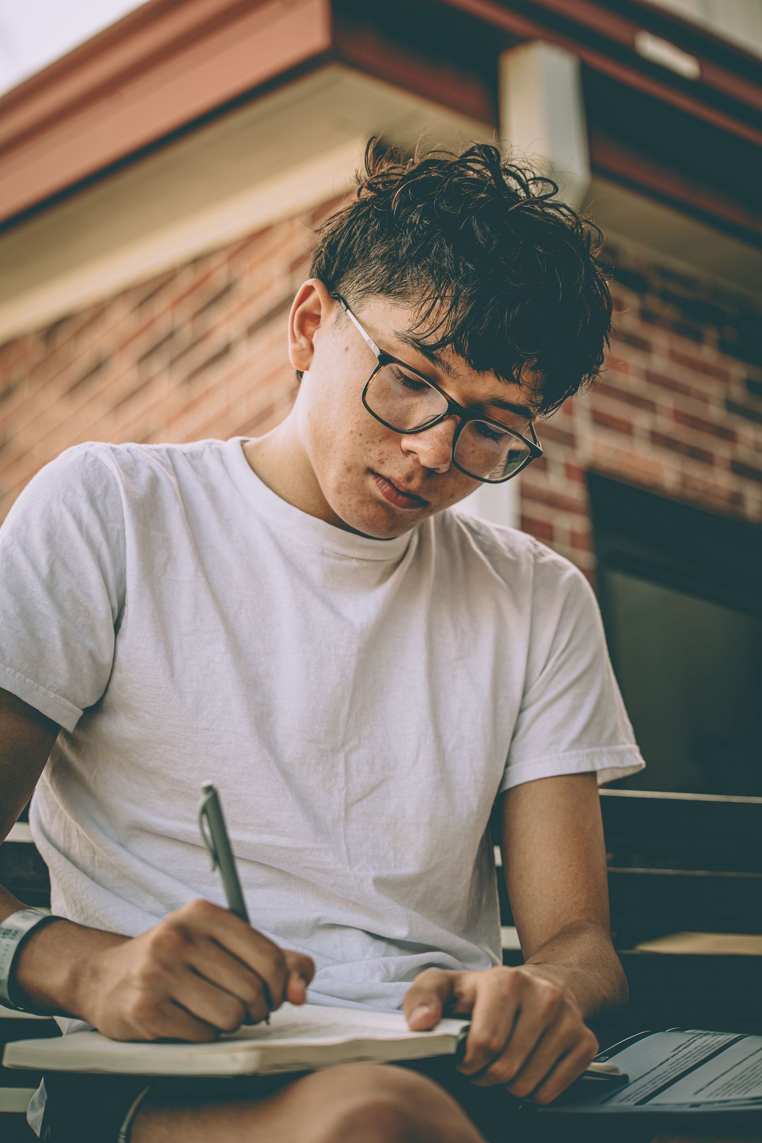 Young man with glasses writing in notebook outdoors near brick building with laptop nearby.