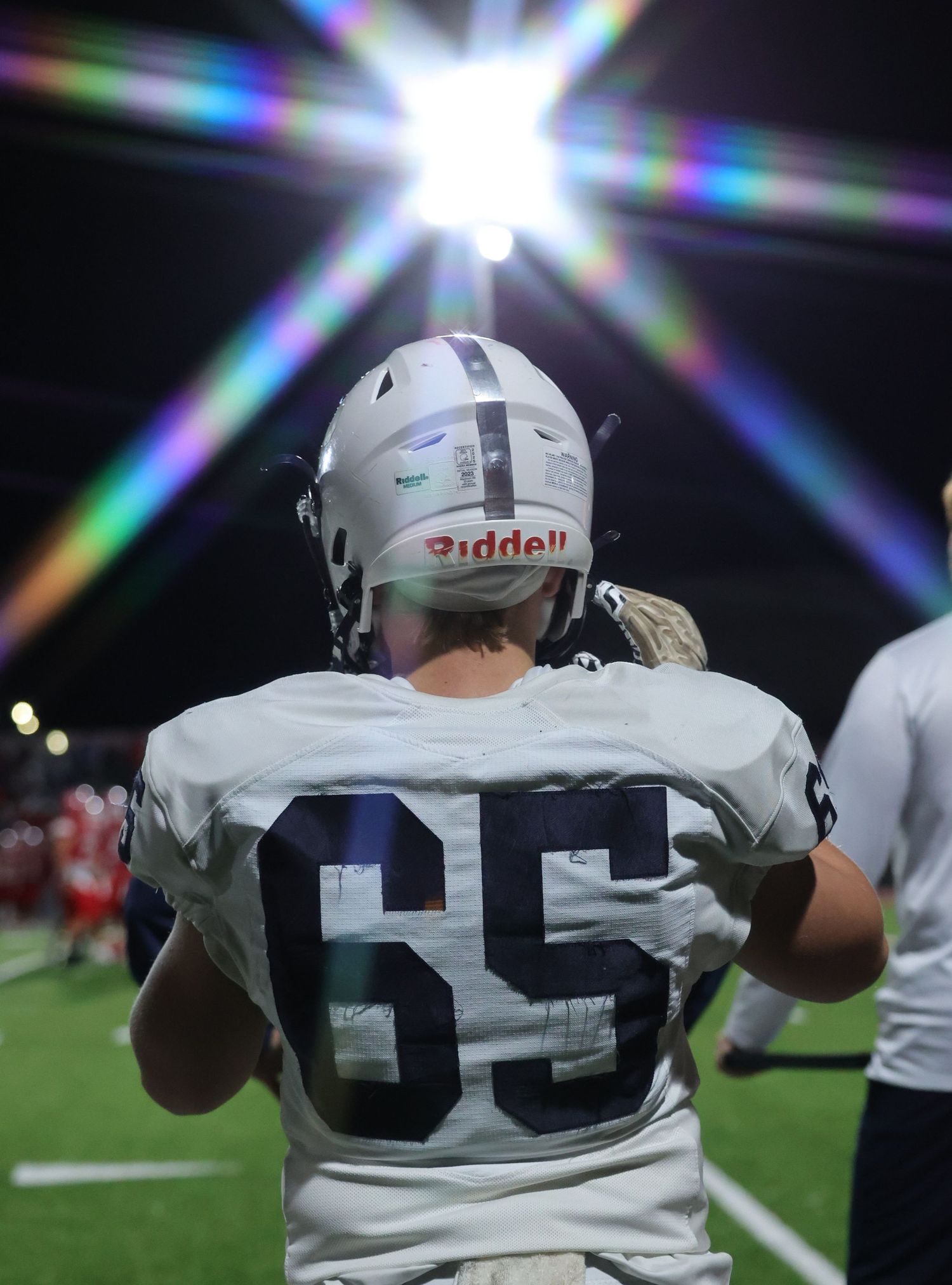 Football player wearing Riddell helmet and number 65 jersey stands on sideline under bright stadium lights.