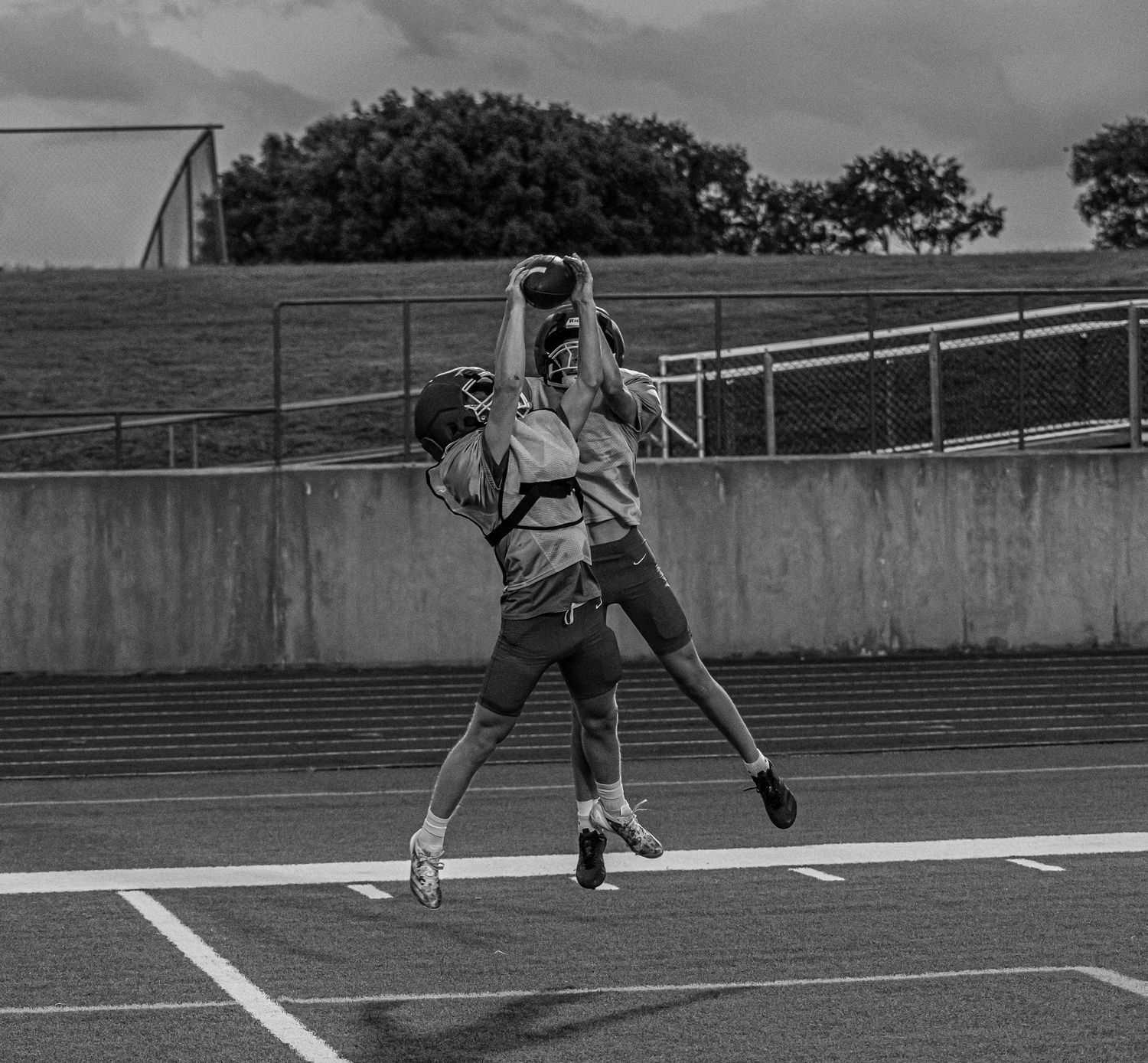 Two athletes jump to catch a football during an outdoor practice session in black and white.