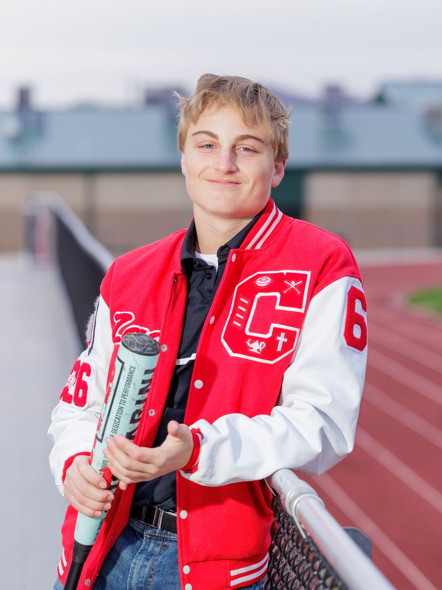 A smiling teen in a red letterman jacket holds a diploma tube while leaning on a stadium railing.