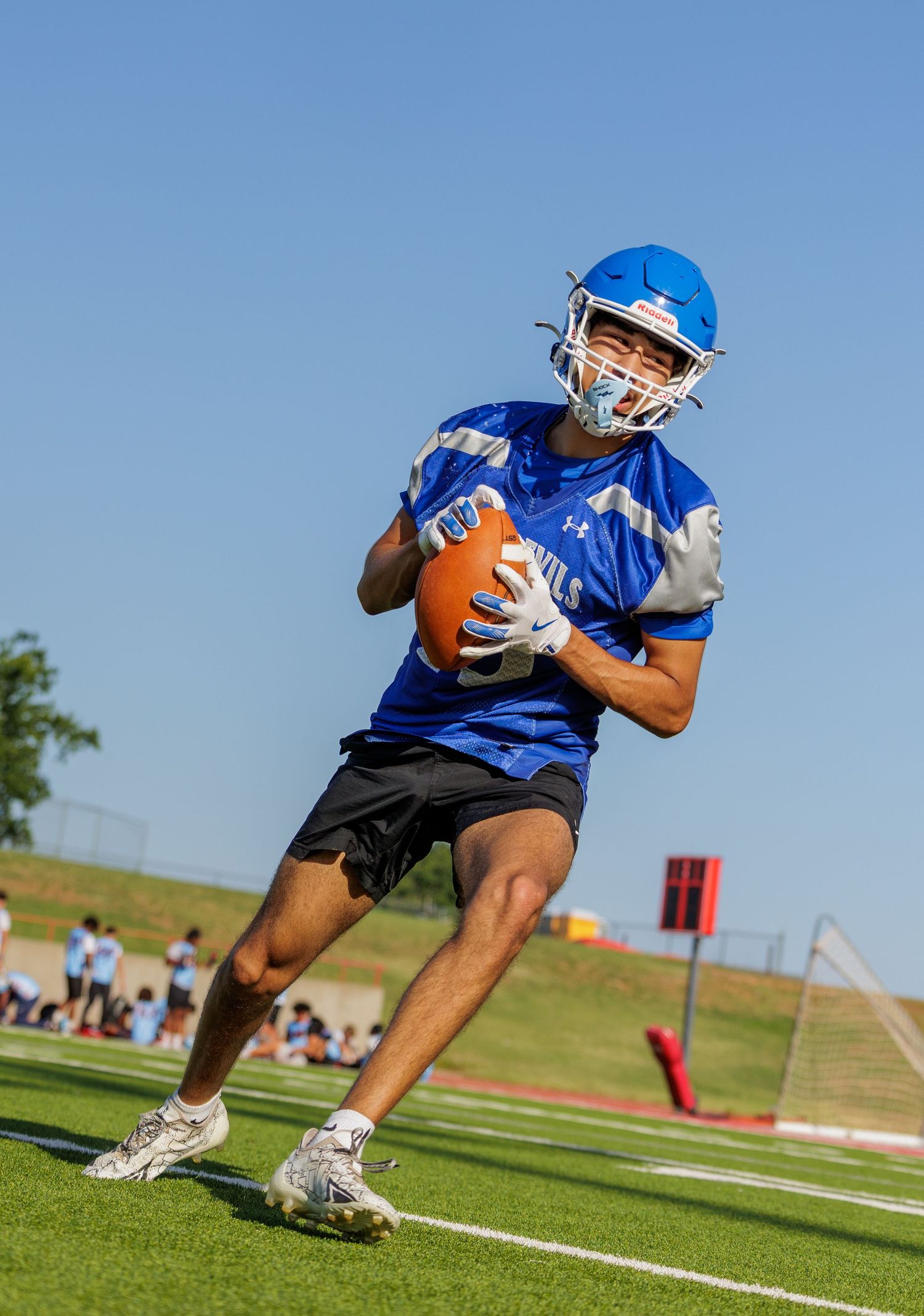 Young football player in blue jersey and helmet catches a football during an outdoor practice session.