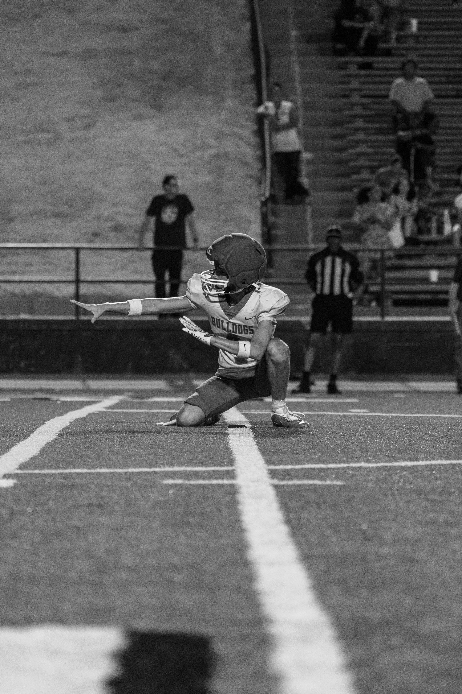 Black and white photo of a football player kneeling on the field with arms outstretched during a game.