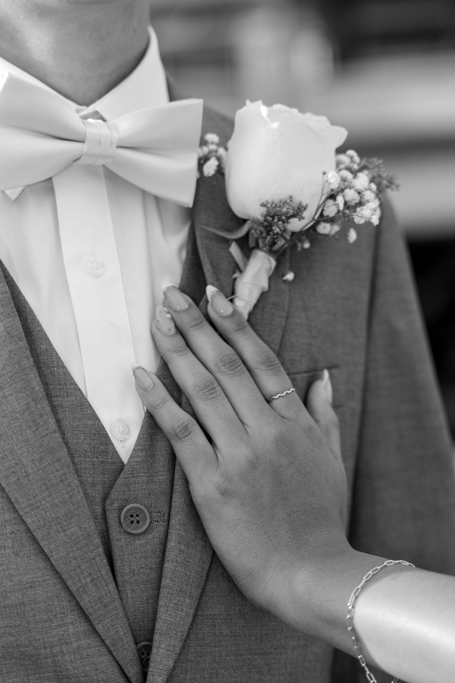 Black and white close-up of a hand adjusting a rose boutonniere on a suit with a bow tie.